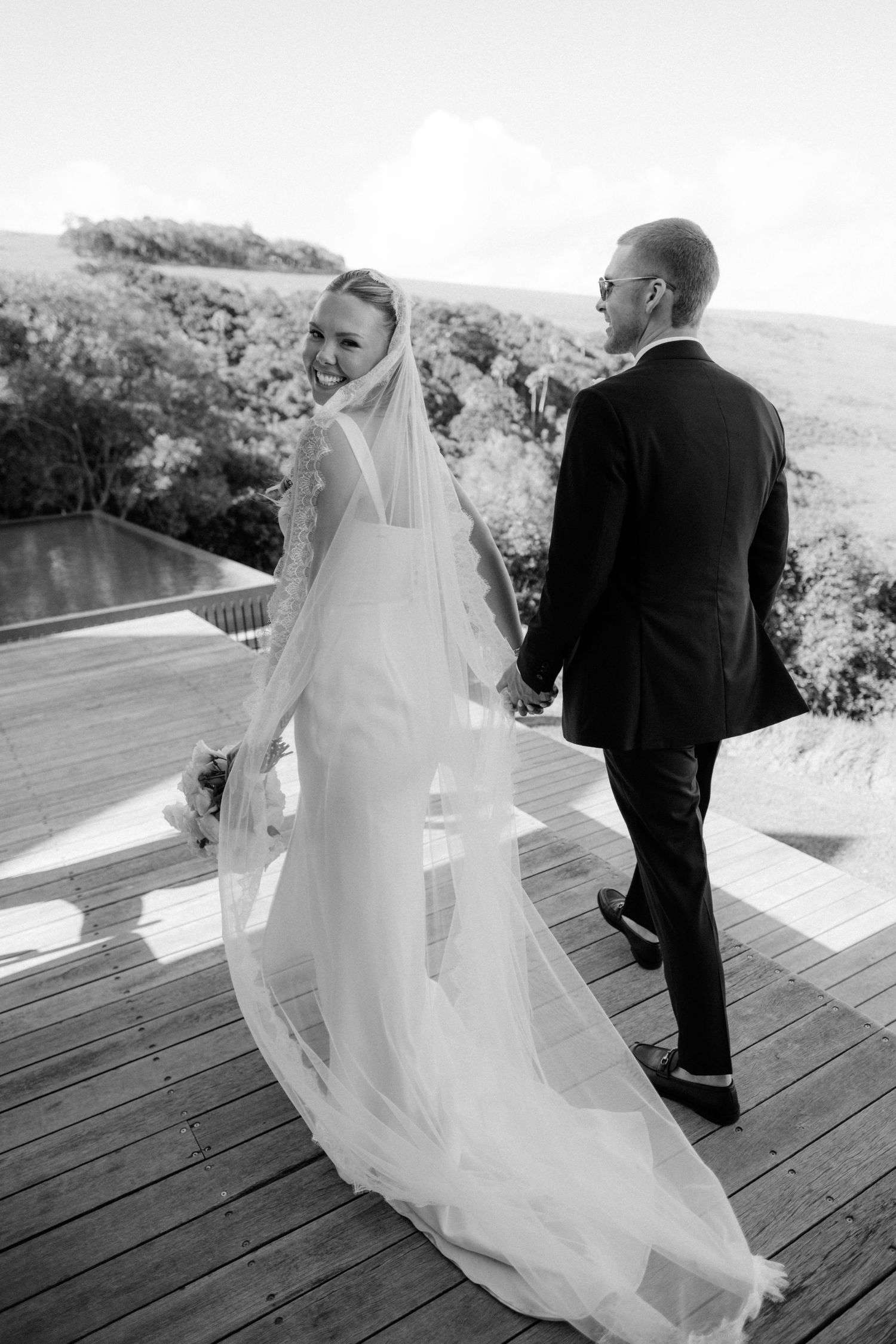 Black and white photo of smiling bride in long veil holding grooms hand walking on outdoor wooden deck.