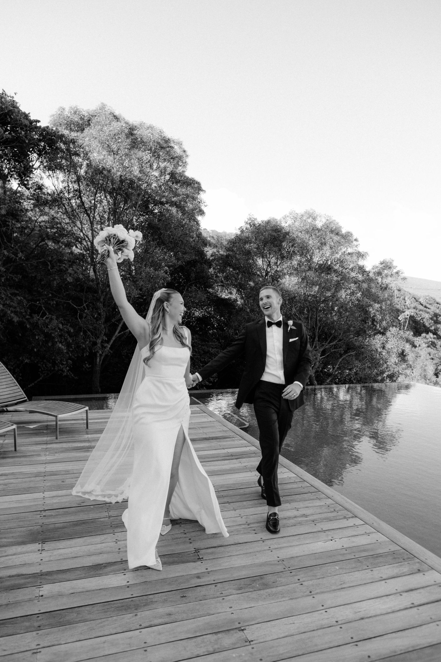 Bride raises bouquet joyfully as groom in tuxedo walks beside her on waterfront deck.