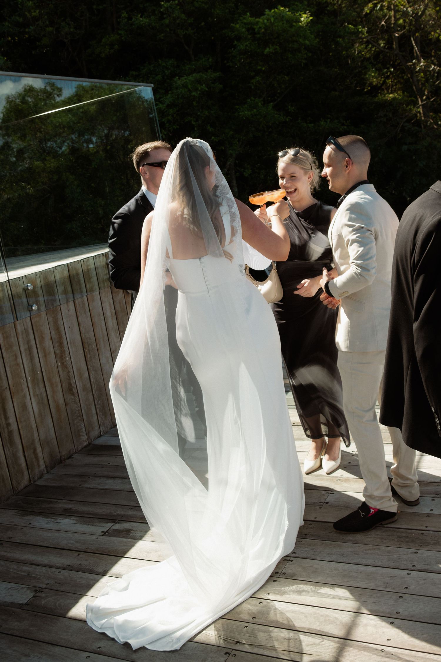 Bride in elegant white gown with flowing veil mingles with guests on a sunlit wooden deck.