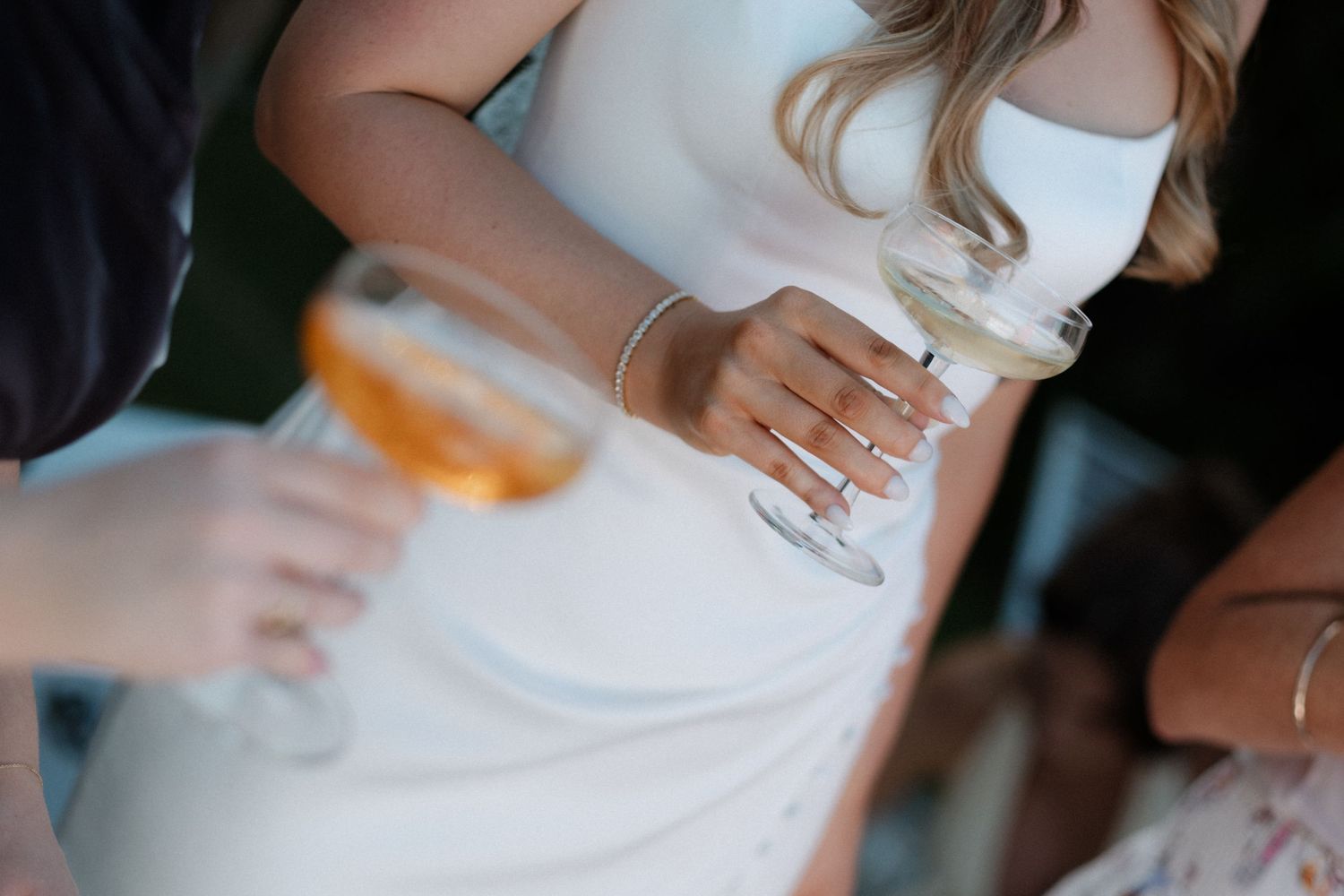 Close-up of bride in white dress holding champagne glass with elegant bracelet and rings.
