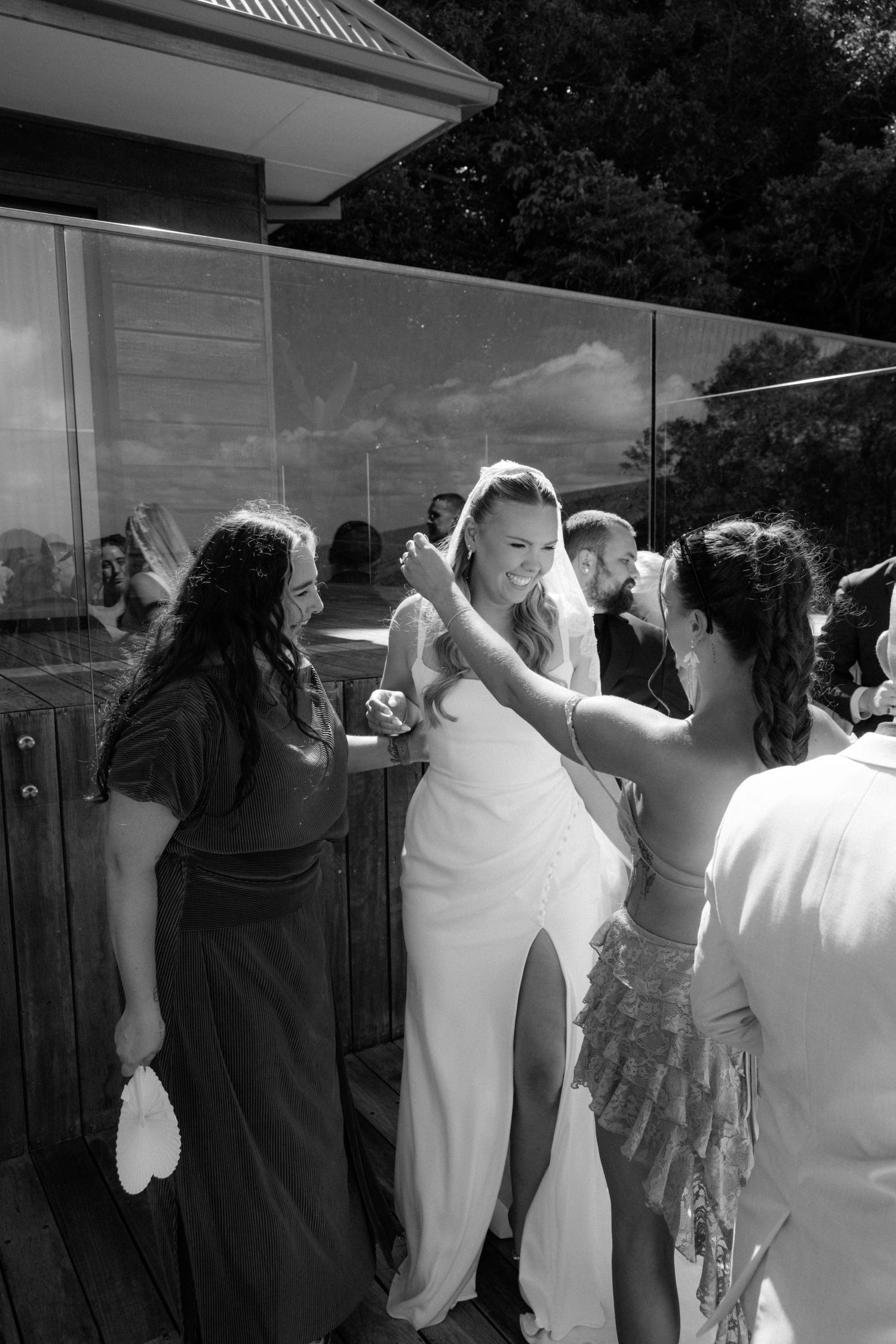 Black and white photo of bride in white gown being assisted by bridesmaids on outdoor deck at wedding.