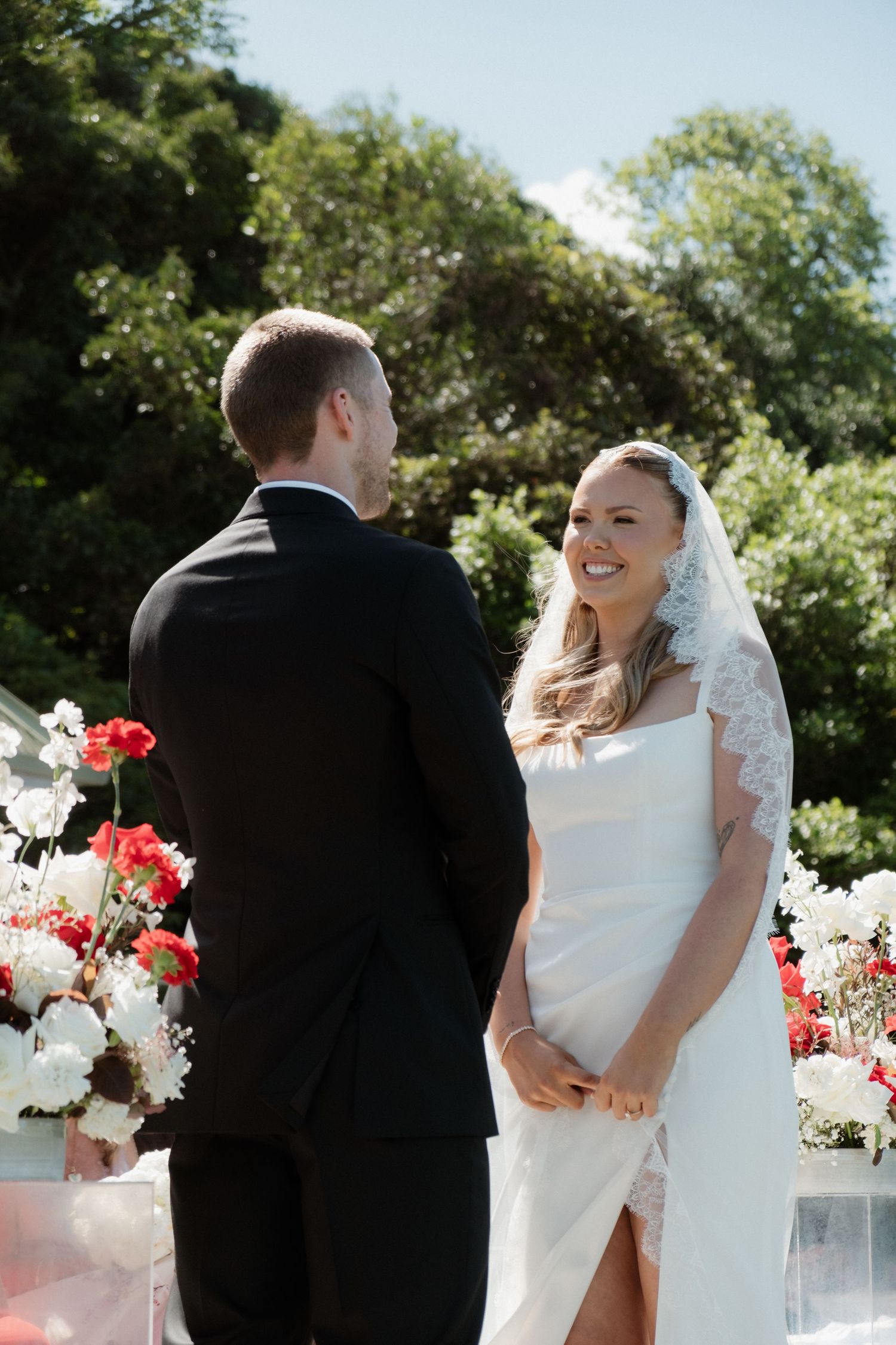 Smiling bride in white gown and lace veil exchanging vows with groom at outdoor floral ceremony.