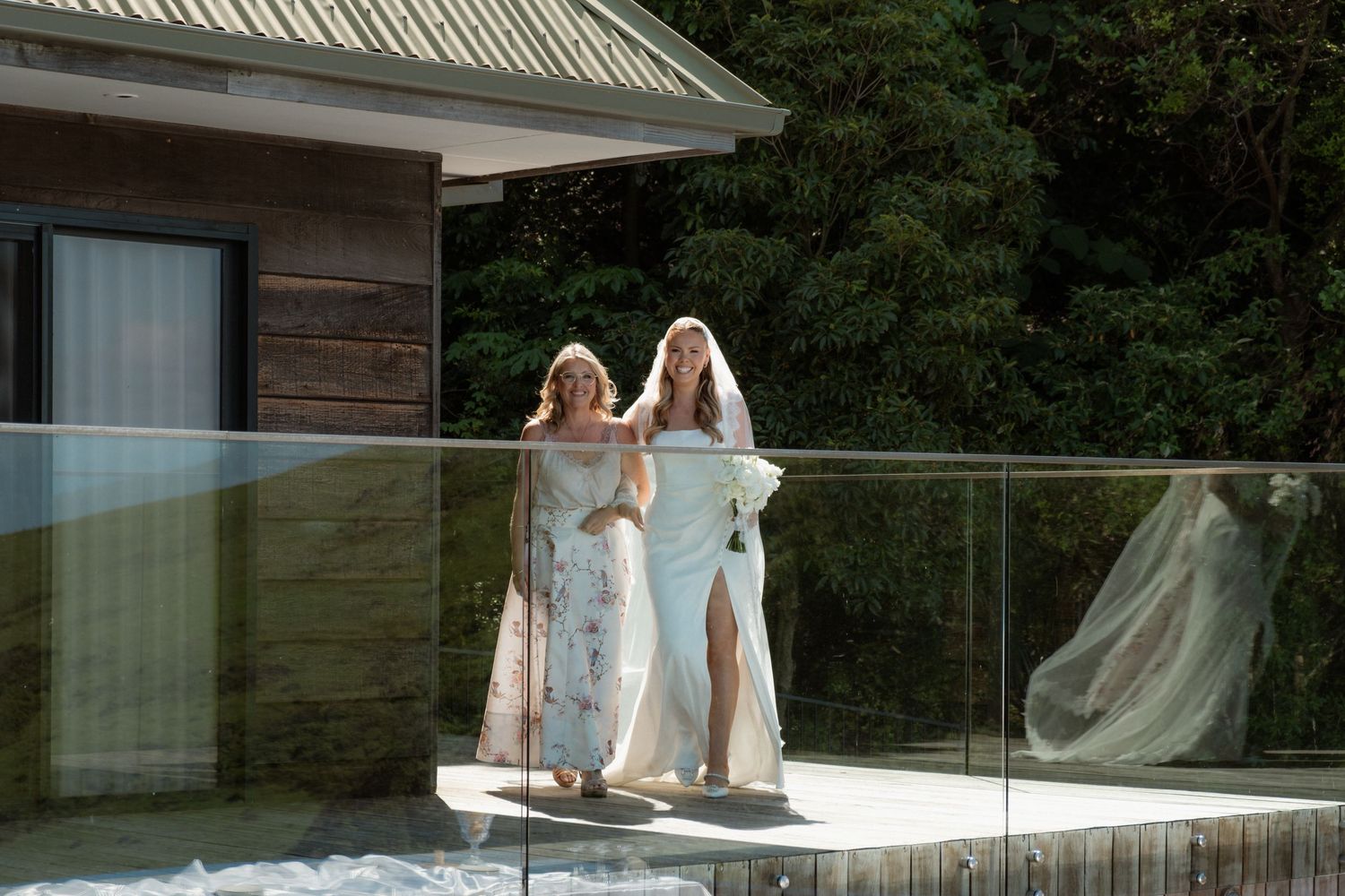 Two women in elegant white dresses stand together on a glass-railed balcony surrounded by lush greenery.