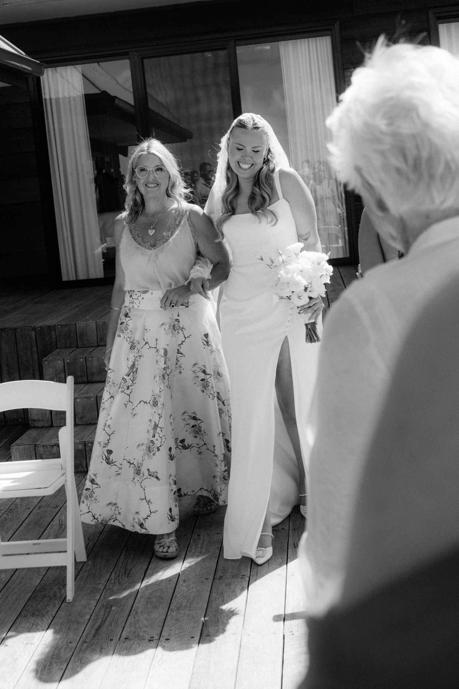 Black and white photo of a smiling bride walking down the aisle with a woman on a sunny deck.