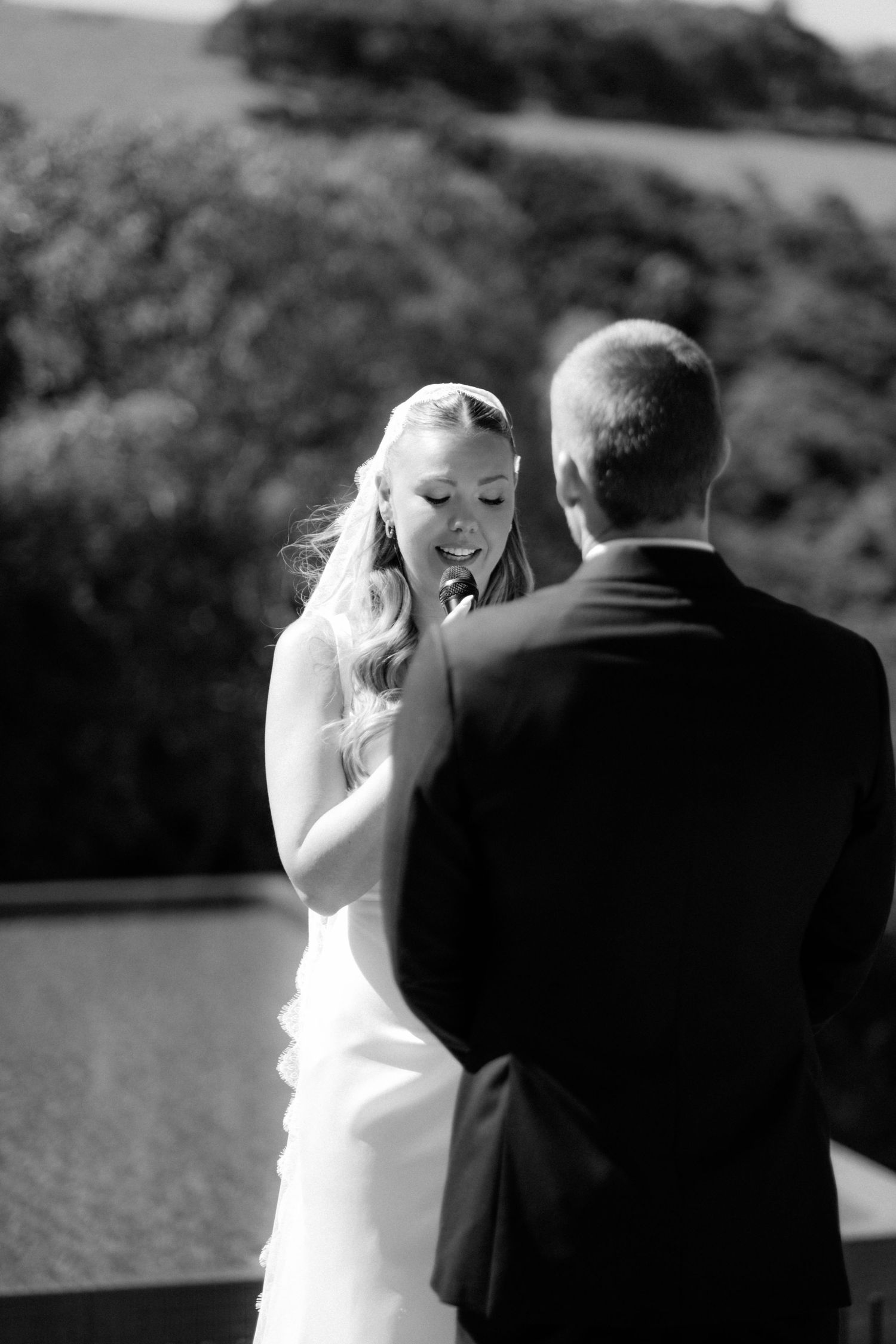 Black and white photo of emotional bride exchanging vows with groom during outdoor wedding ceremony.