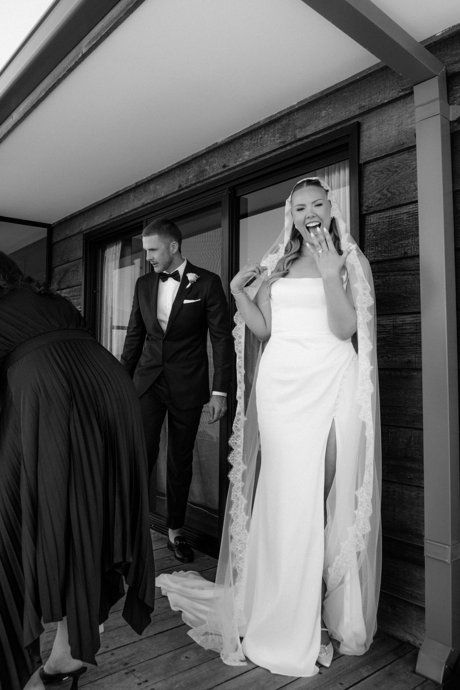 Black and white photo of a bride in a strapless gown with veil and groom in tuxedo on a wooden deck.