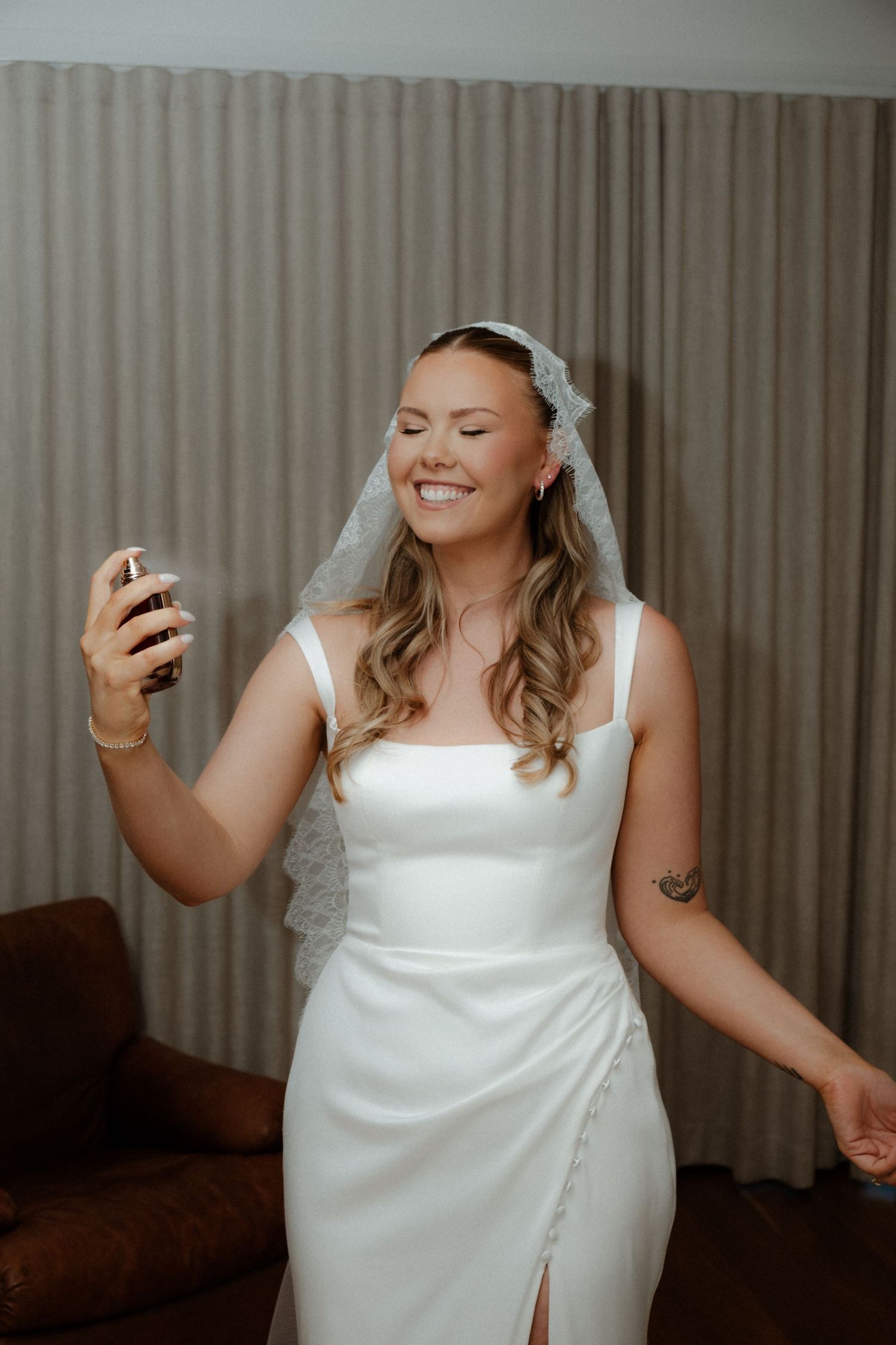 Smiling bride in white square-neck wedding dress and lace veil applying perfume indoors.