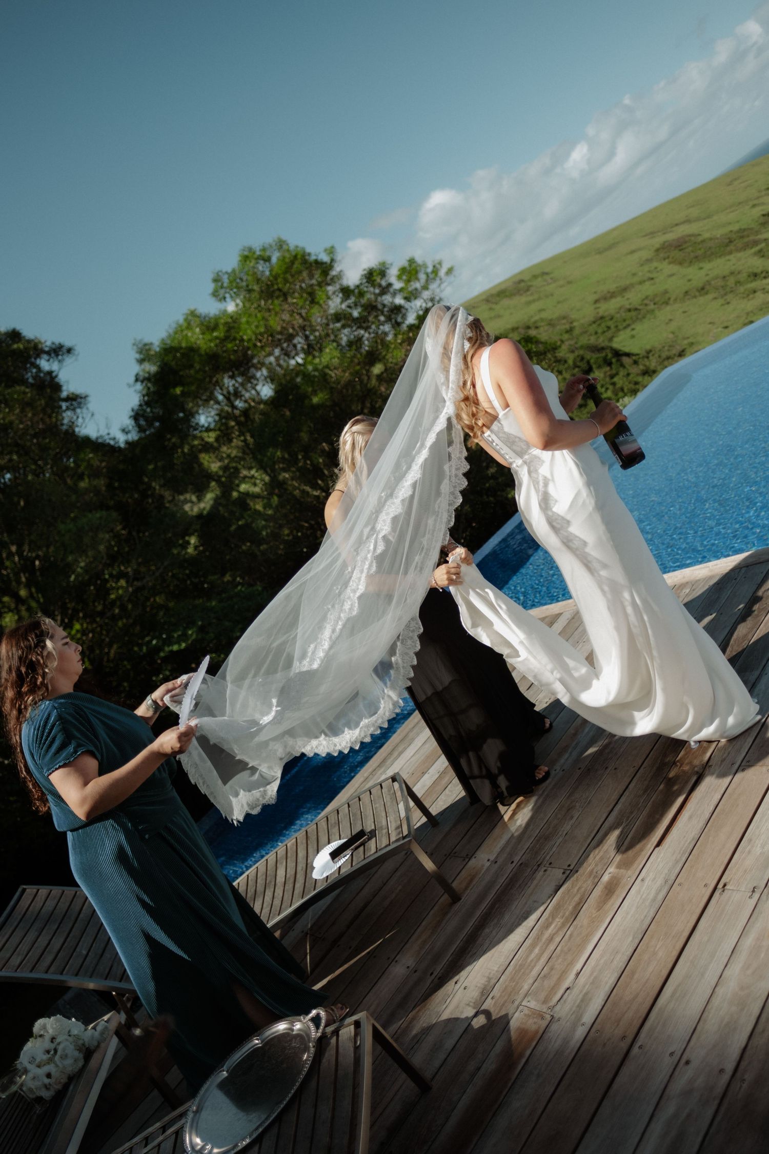 A bridesmaid helps adjust the brides flowing veil on a sunny wooden deck beside a pool with green hills.