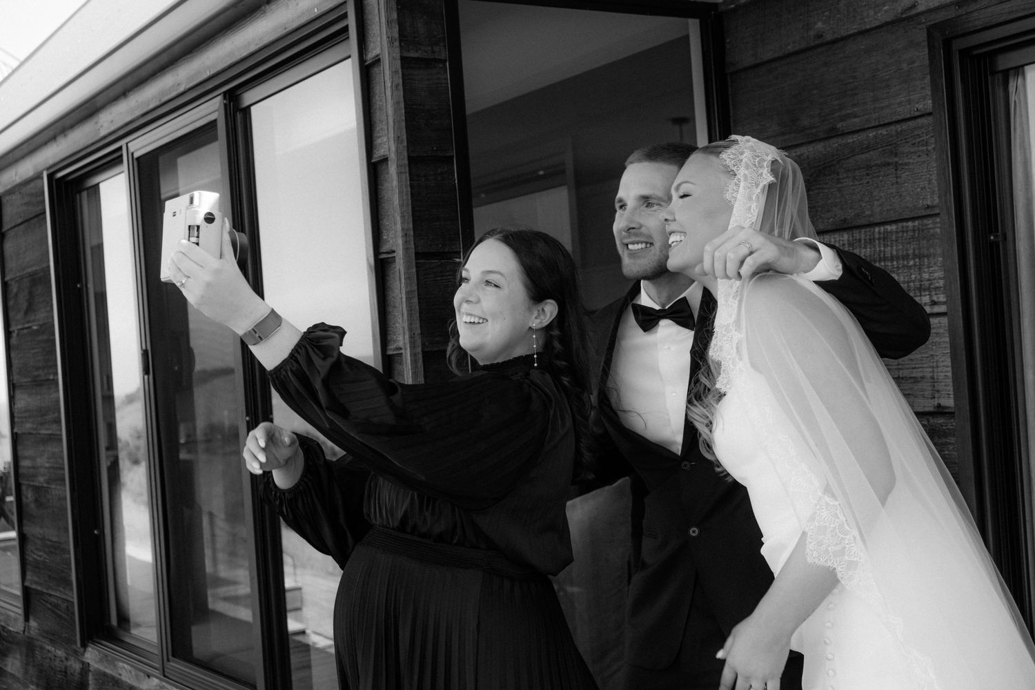 Black and white photo of bride and groom taking a selfie with a guest at a wedding reception.