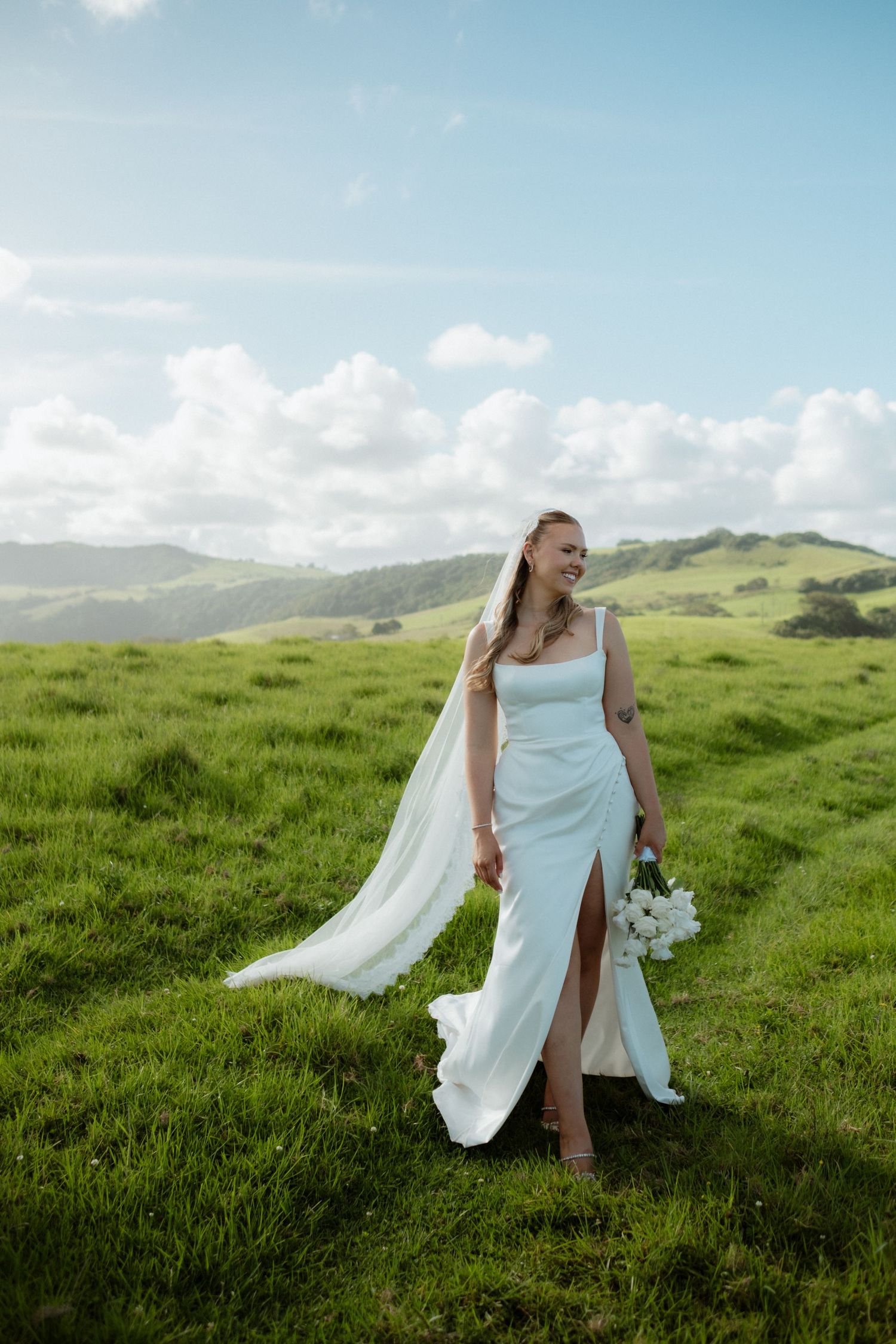 Bride in white gown and veil holding bouquet standing in lush green hillside landscape.