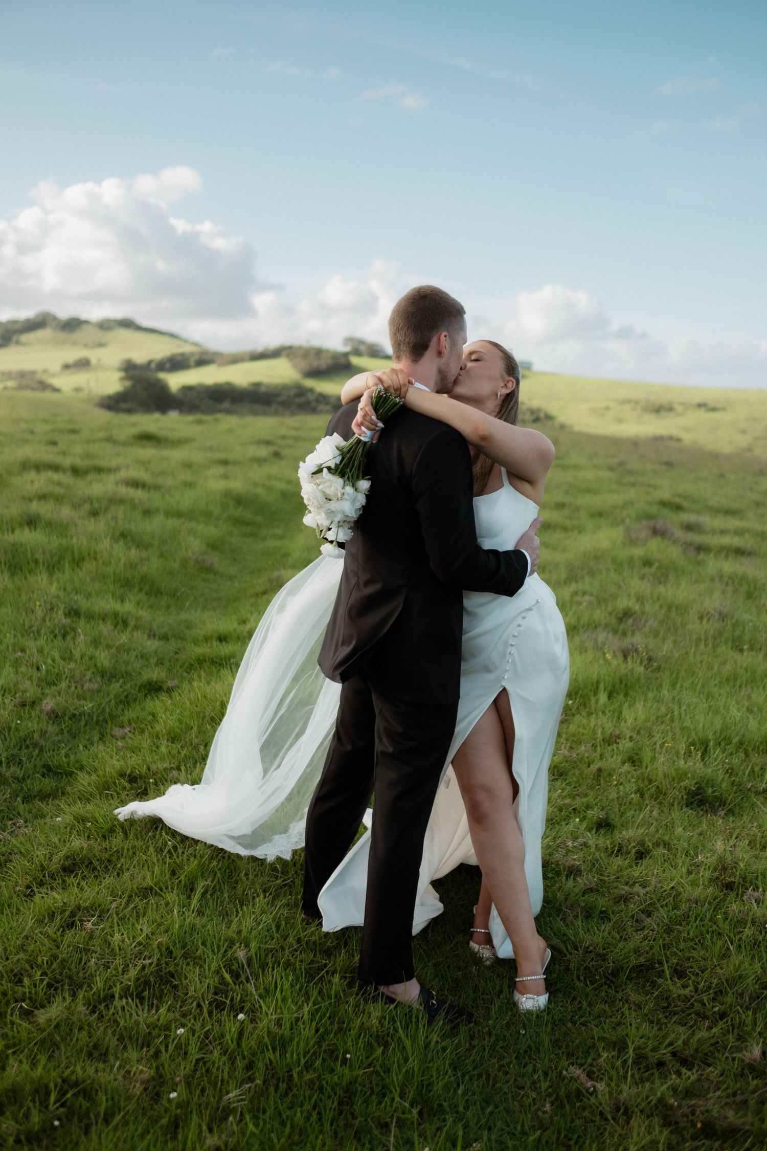 Bride and groom kissing in lush green meadow, bride holding white bouquet with veil flowing.