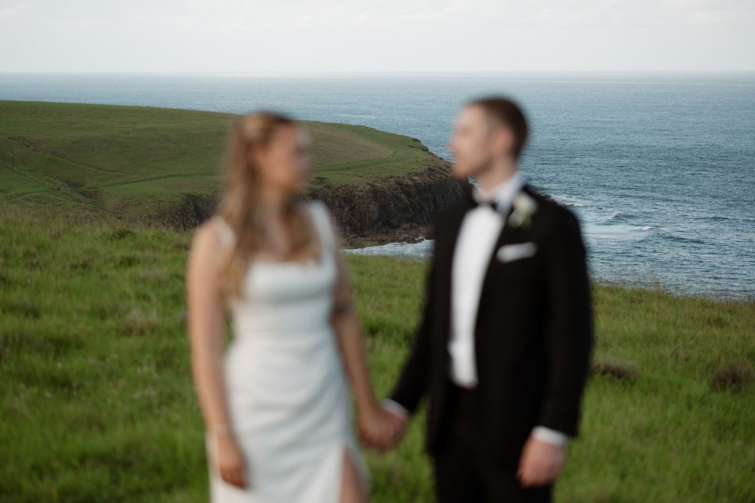 Bride and groom holding hands on lush green coastal cliffs with ocean views on their wedding day.