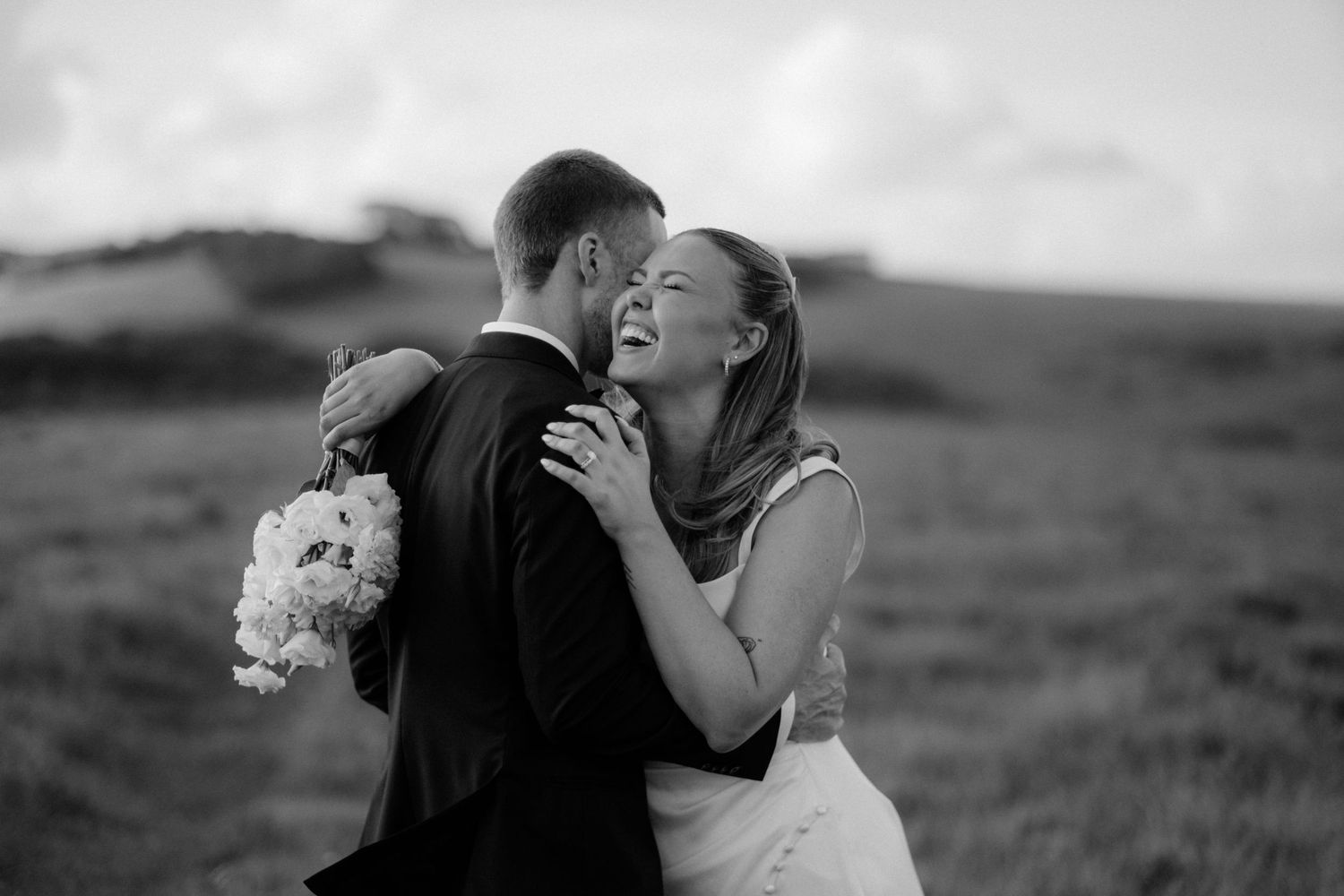 Black and white photo of a laughing bride embracing a groom in a field, holding a white bouquet.