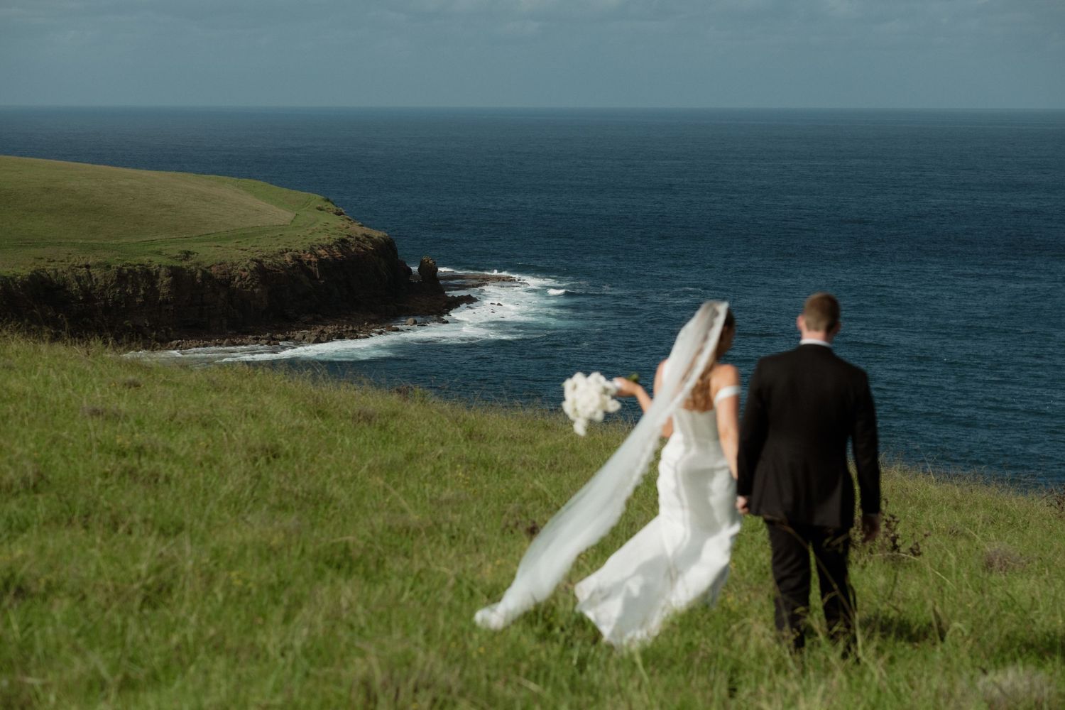 Bride and groom walking on a green coastal cliff overlooking the ocean on their wedding day.