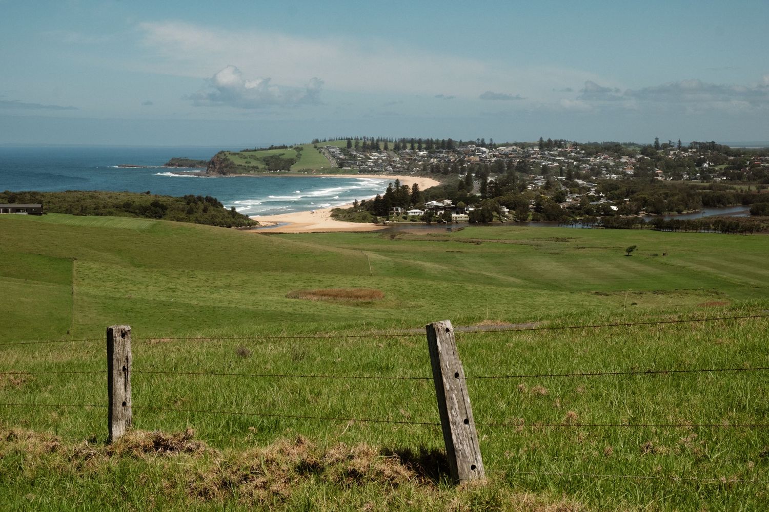 Scenic coastal landscape with green farmland, fence posts, sandy beach and ocean town beyond.