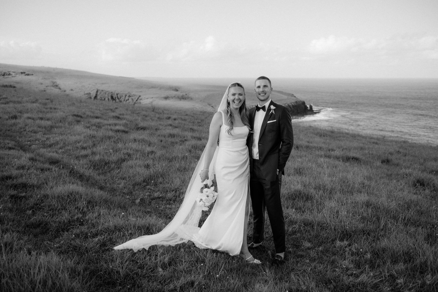 A smiling bride and groom pose together on a grassy coastal hillside in a classic black and white portrait.
