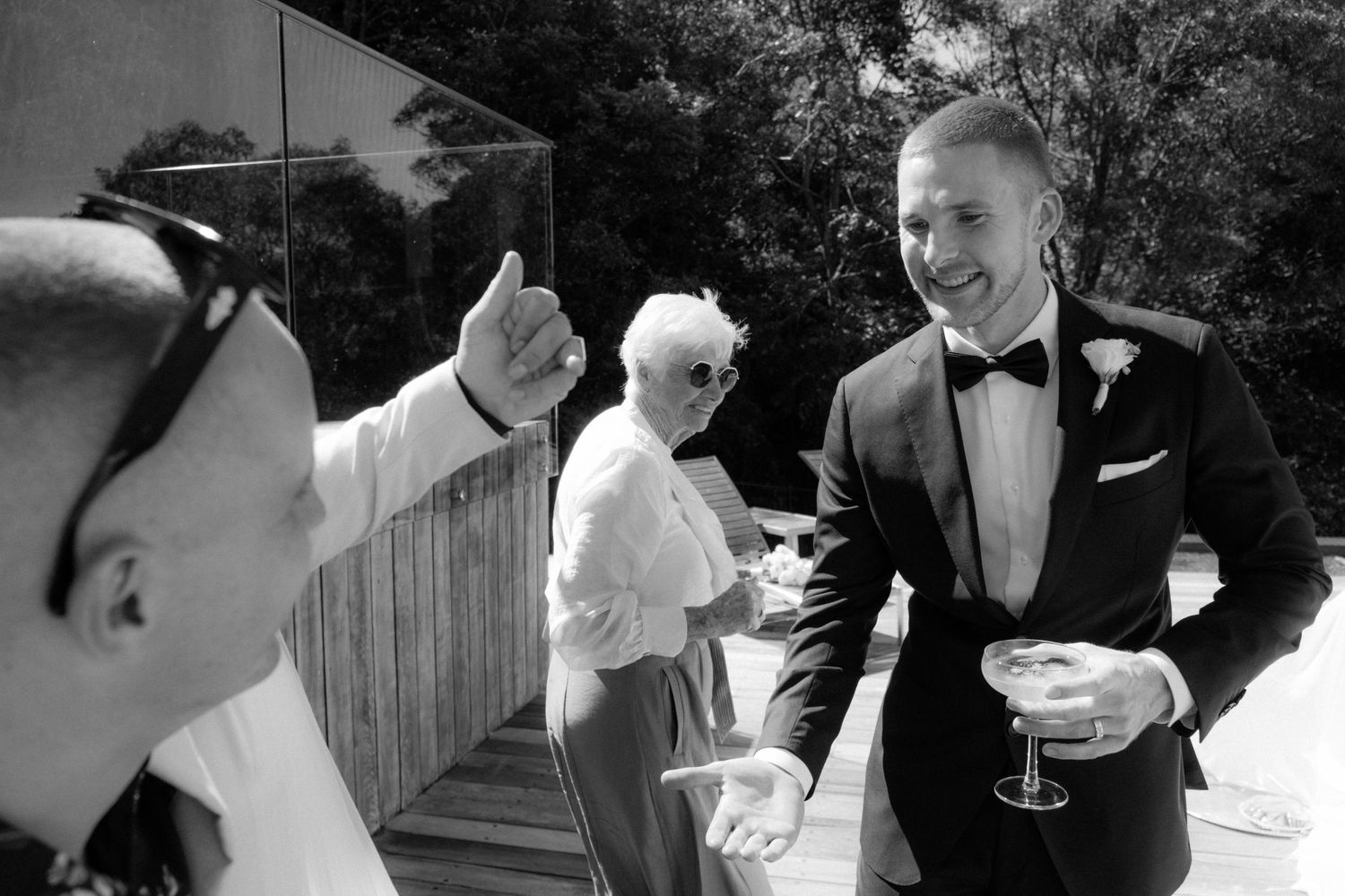 Groom in black tuxedo smiling with guests holding champagne at outdoor wedding reception.
