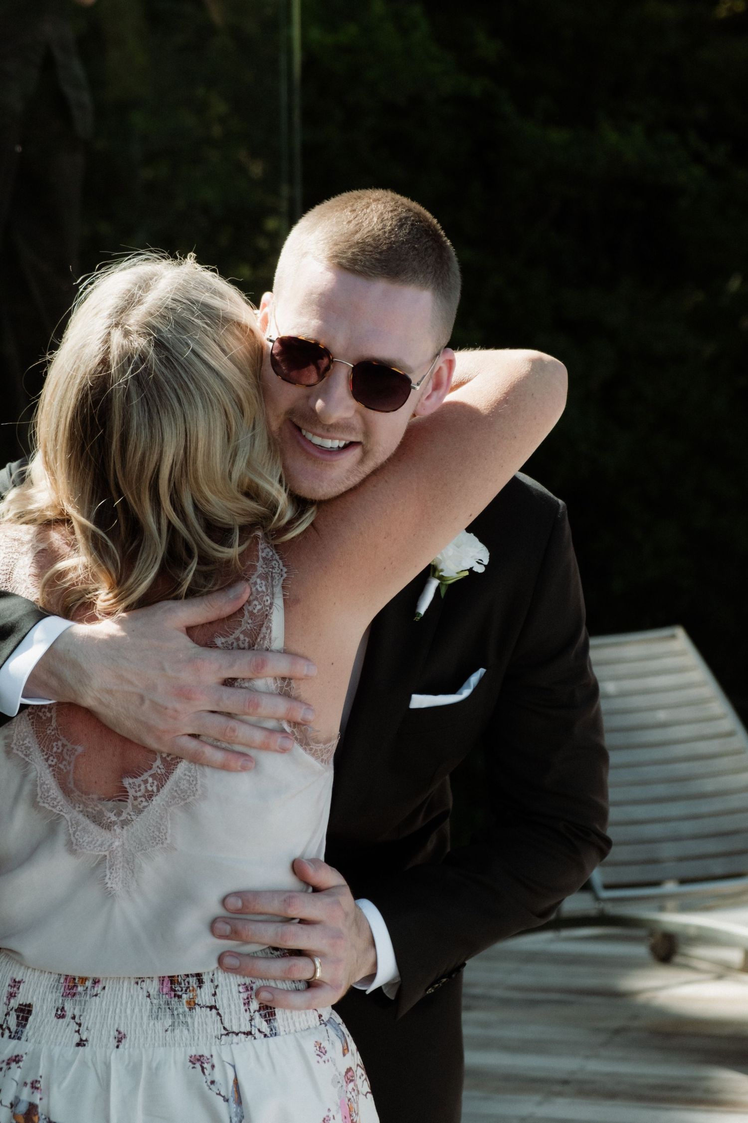 A smiling groom in a black suit and sunglasses shares a warm embrace with a wedding guest outdoors.