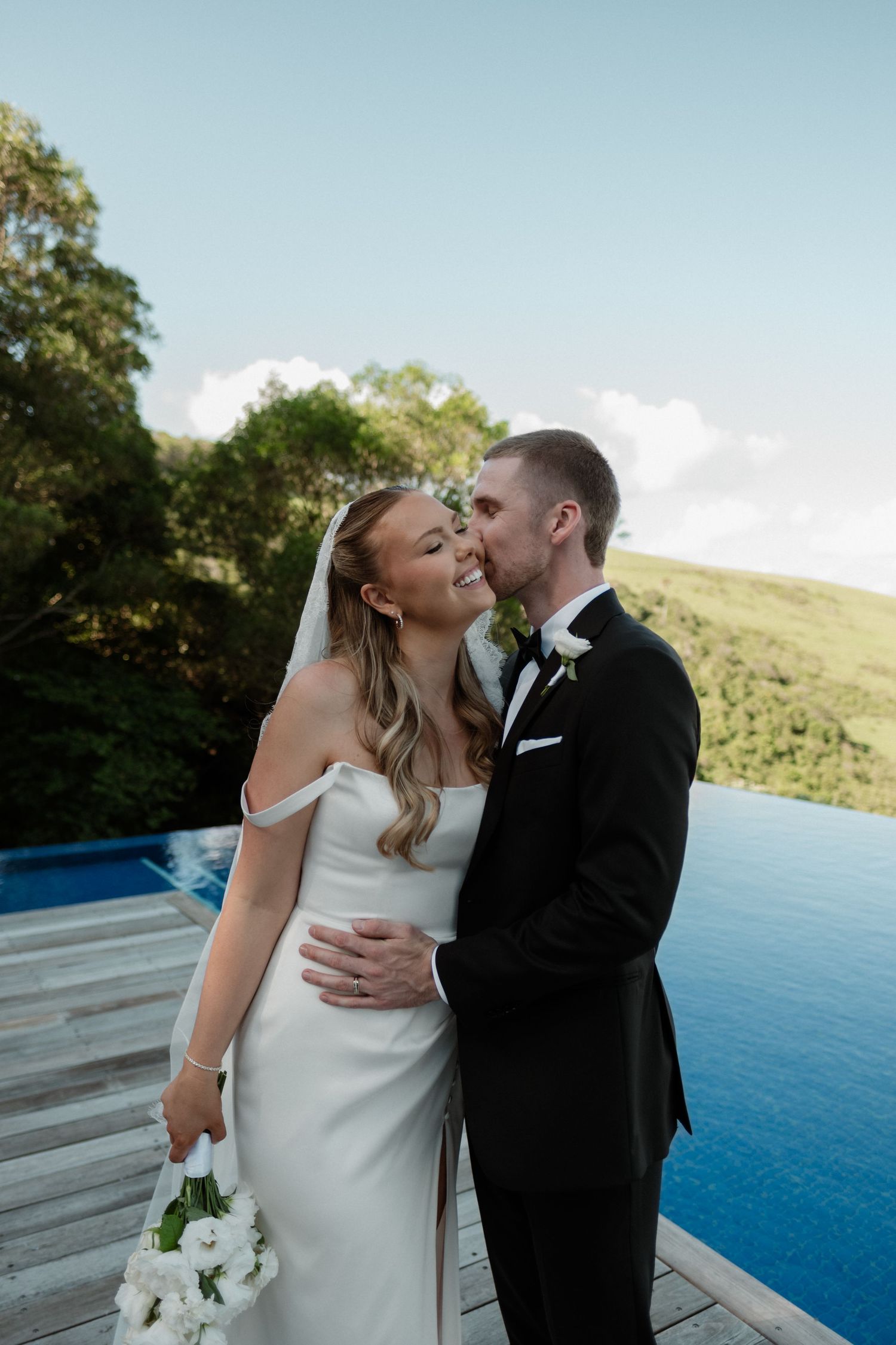 Bride and groom share a romantic moment by an infinity pool surrounded by lush greenery on their wedding day.
