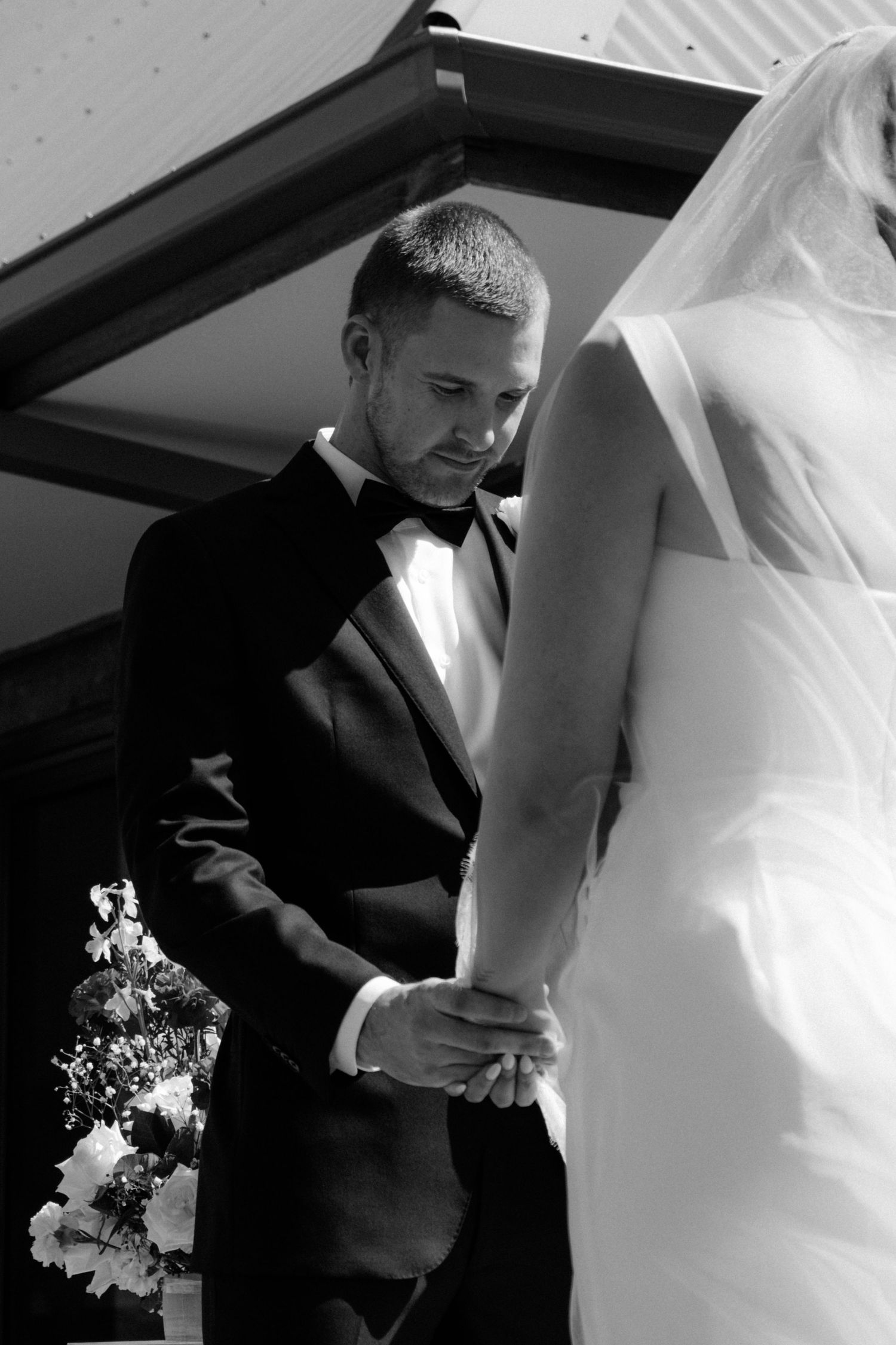 Black and white photo of a groom in a tuxedo holding his bride's hand during a wedding ceremony.