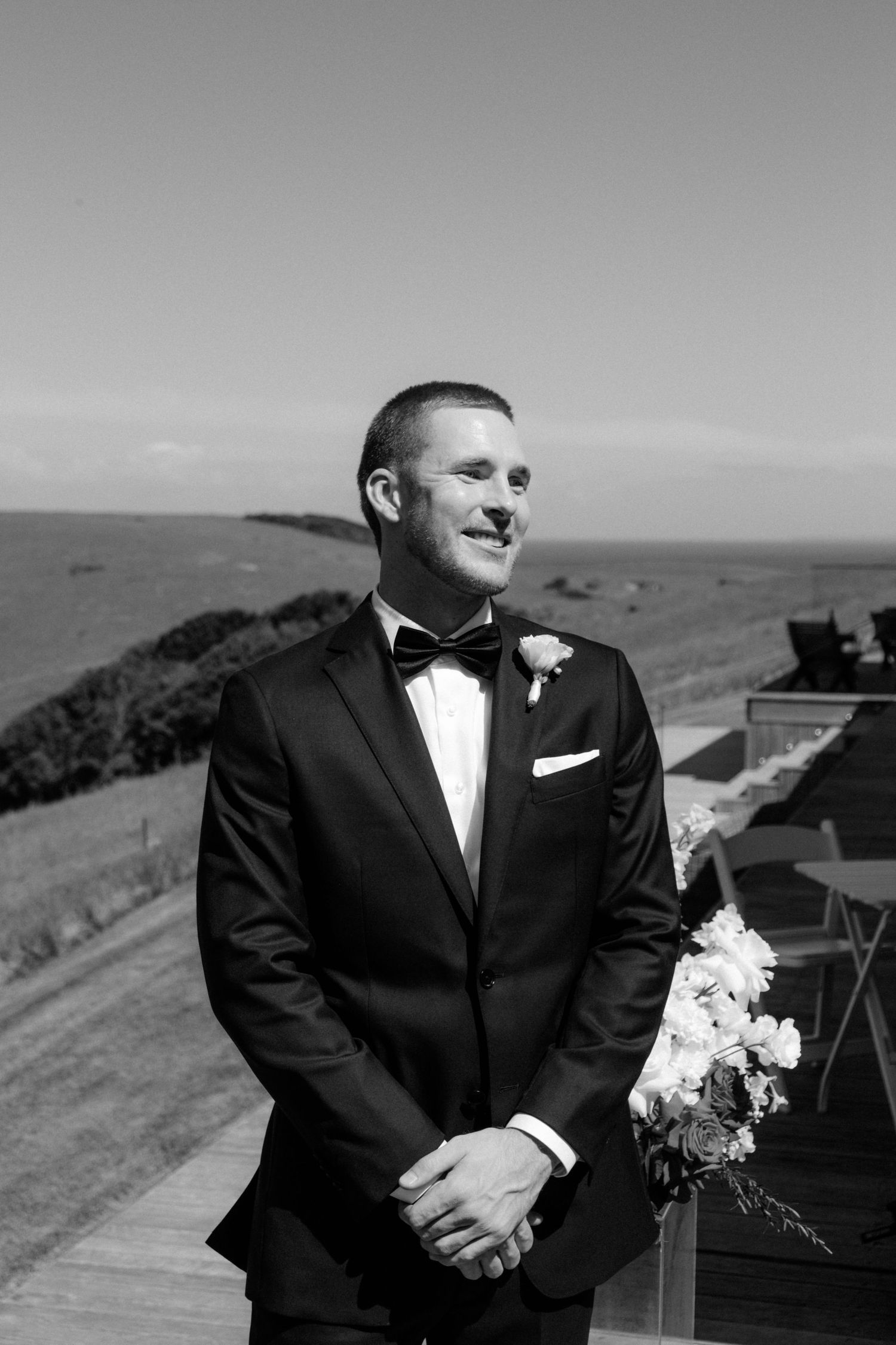 Black and white portrait of a dapper groom in tuxedo and bow tie smiling outdoors by the sea.