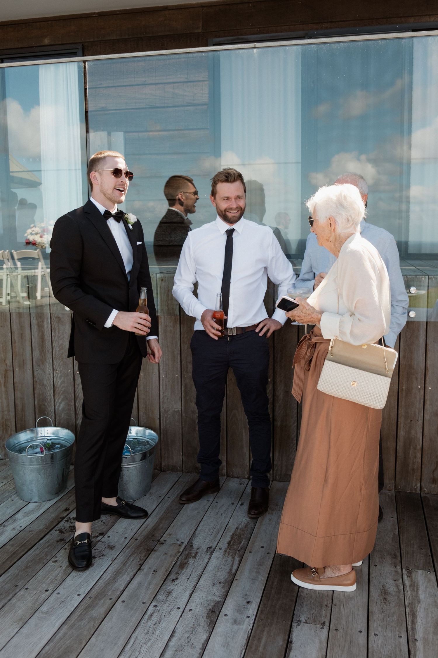 A groom in a tuxedo laughs with two guests on a wooden deck during a sunny outdoor wedding reception.