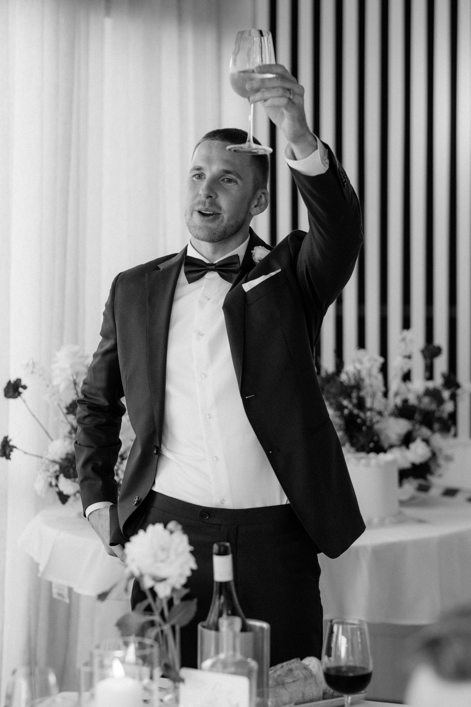 Groom in black tuxedo raises champagne glass for a toast at elegant wedding reception.