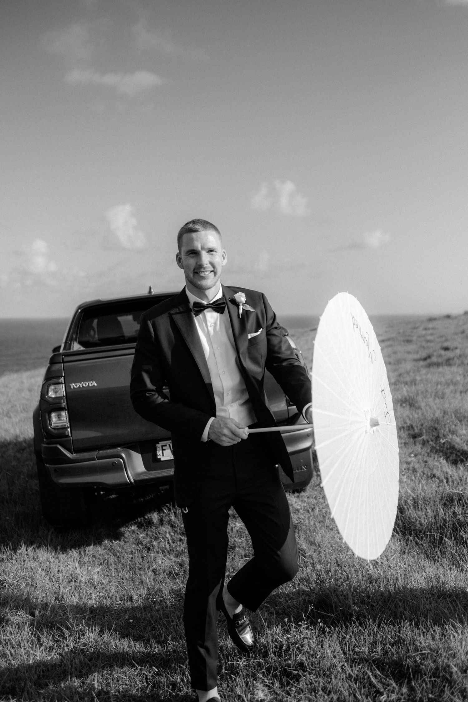 Smiling groom in tuxedo holds white parasol beside Toyota SUV on coastal grassy hillside.