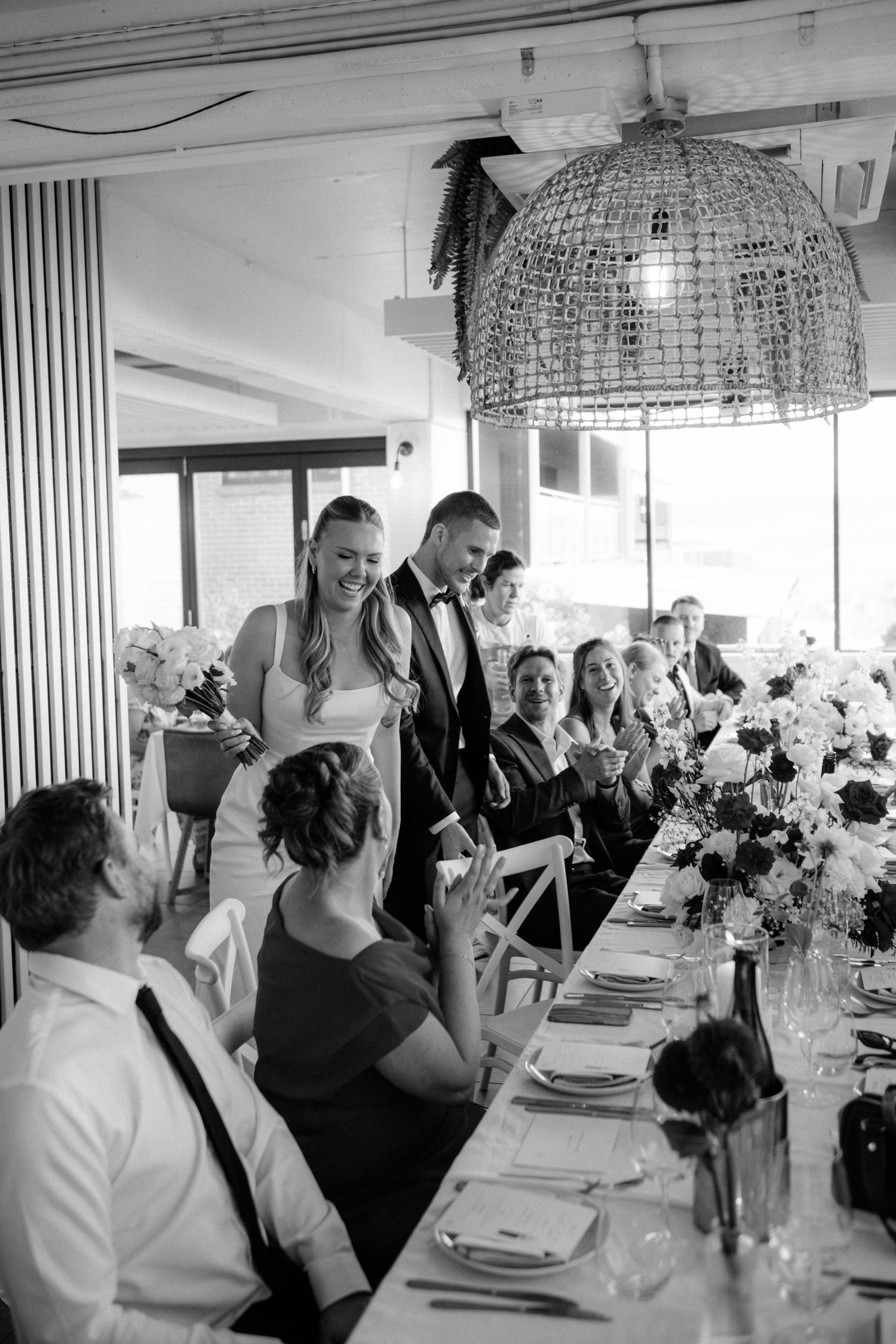 Black and white photo of smiling bride and groom greeting guests at wedding reception dinner table.