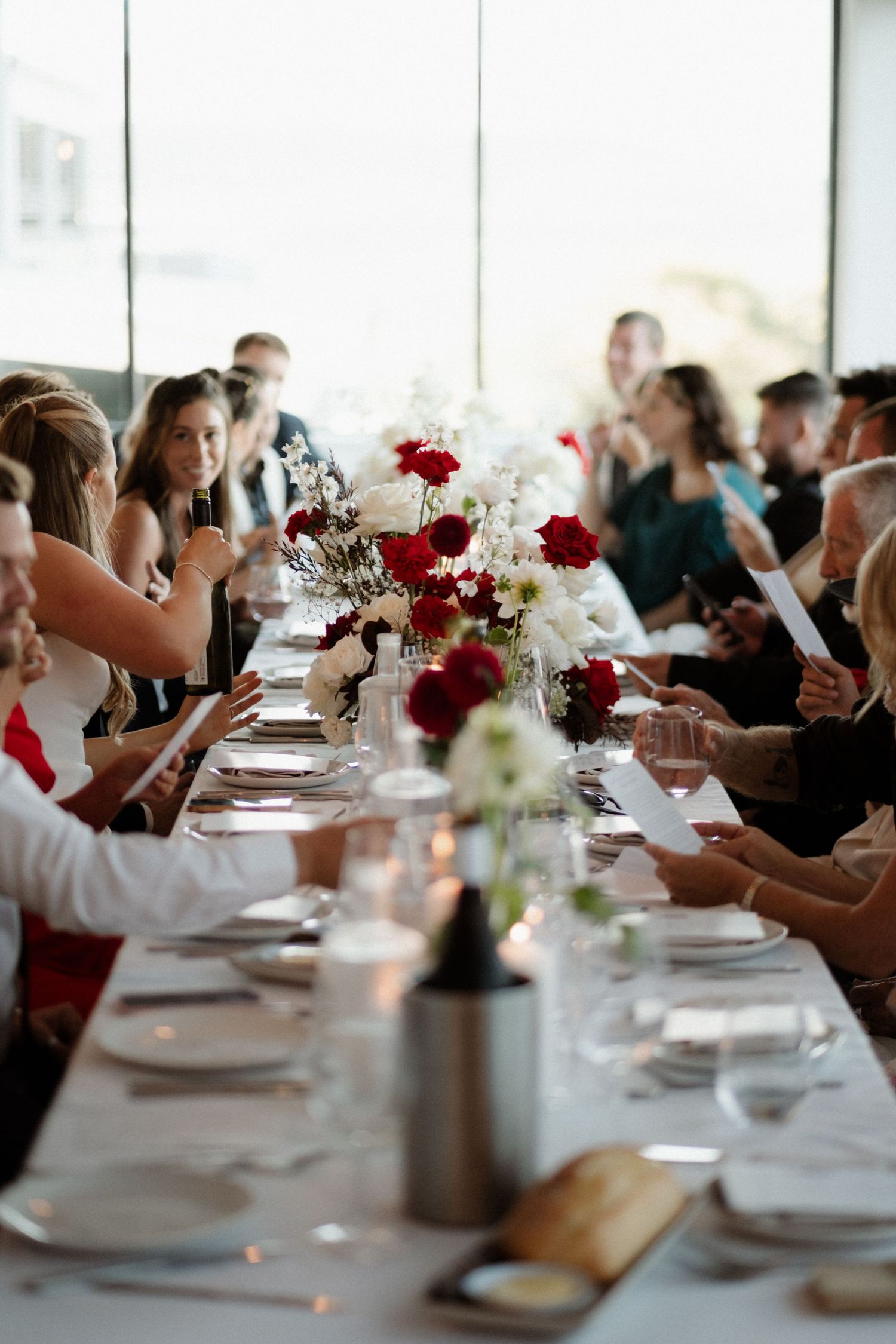 Wedding reception guests seated at a long decorated table with red and white floral centerpieces.