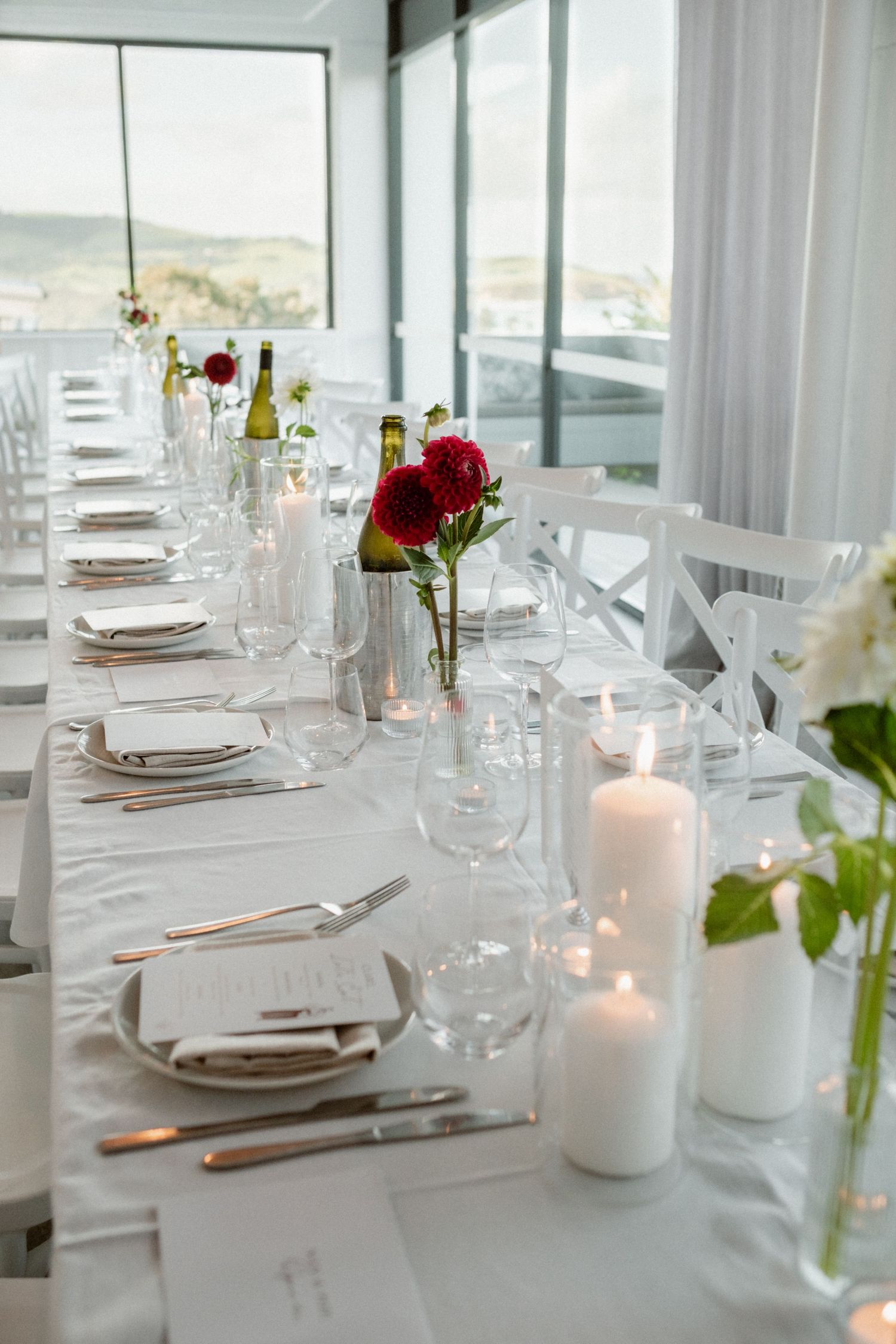 Elegant wedding reception table set with white linens, red roses, candles, and glassware with ocean views.