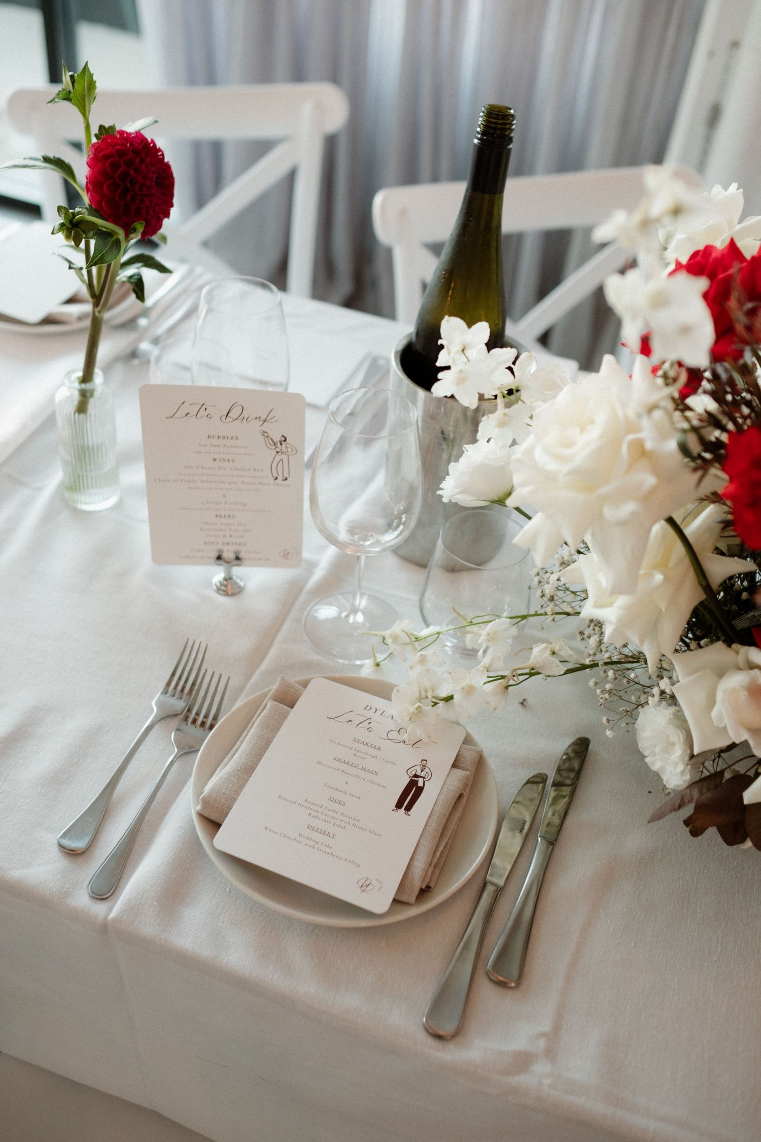 Elegant wedding reception table set with white linens, red and white floral arrangements, and personalized menus.