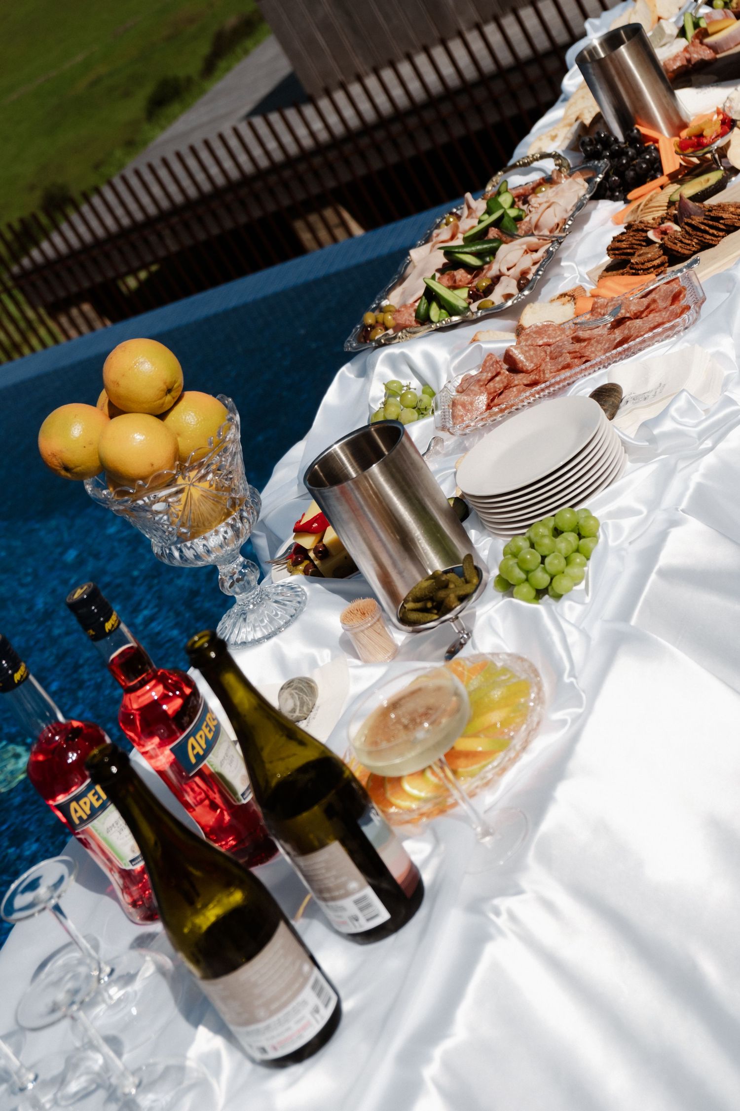 Elegant outdoor wedding reception table with white linen, wine bottles, fresh fruit, charcuterie platters, and floral arrangements.