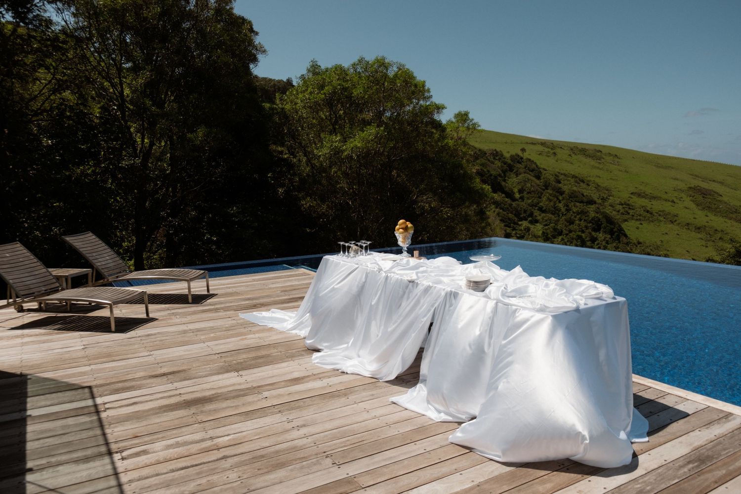 Elegant outdoor dining table with white linen beside an infinity pool on a wooden deck overlooking lush green hills.