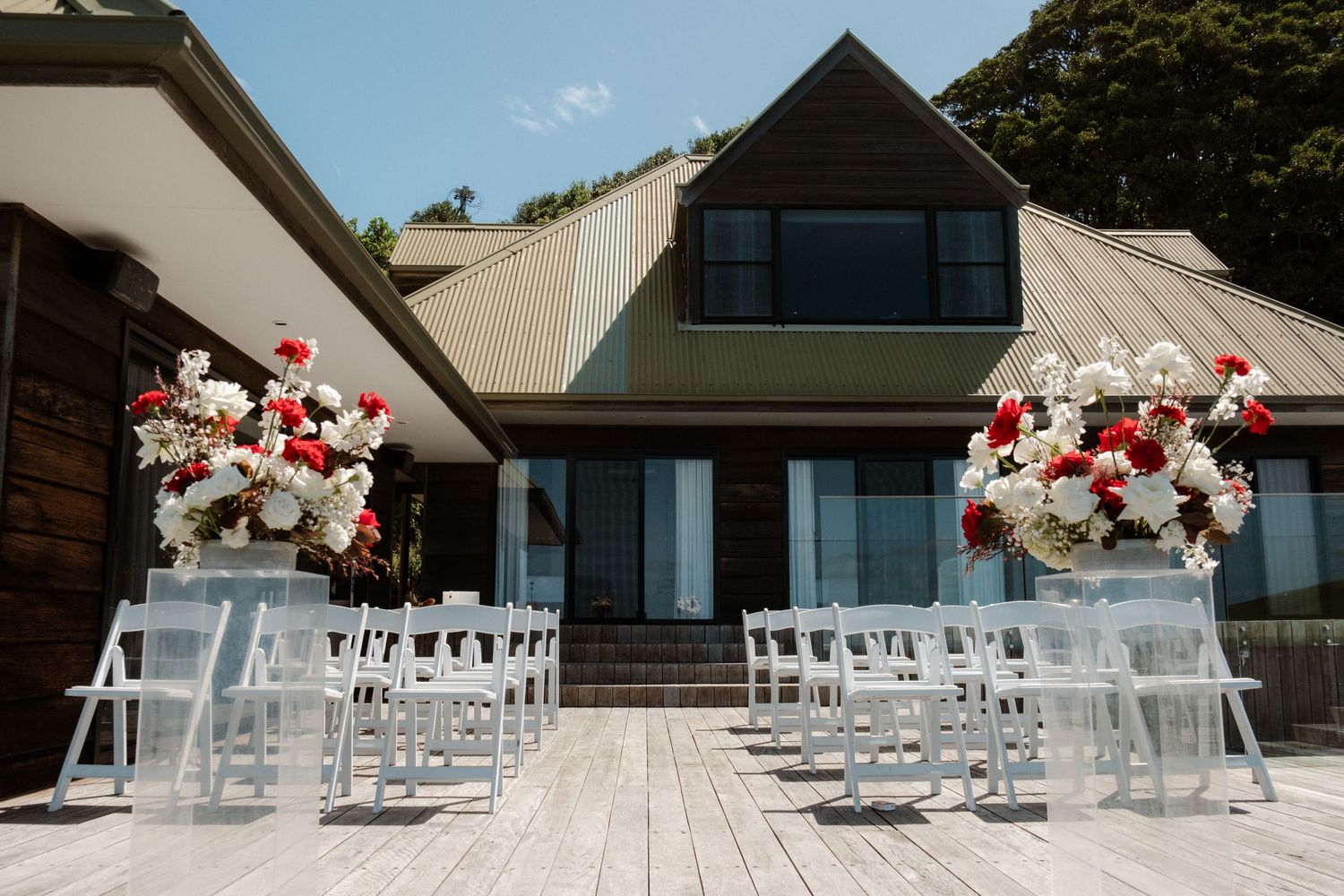 Outdoor wedding ceremony setup with white chairs and red and white floral arrangements on a wooden deck.