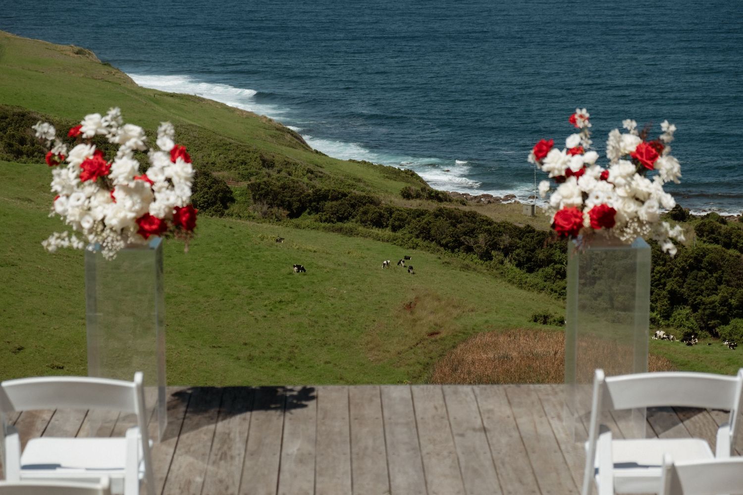 Outdoor wedding ceremony setup with red and white floral arrangements on a coastal cliff overlooking the ocean.