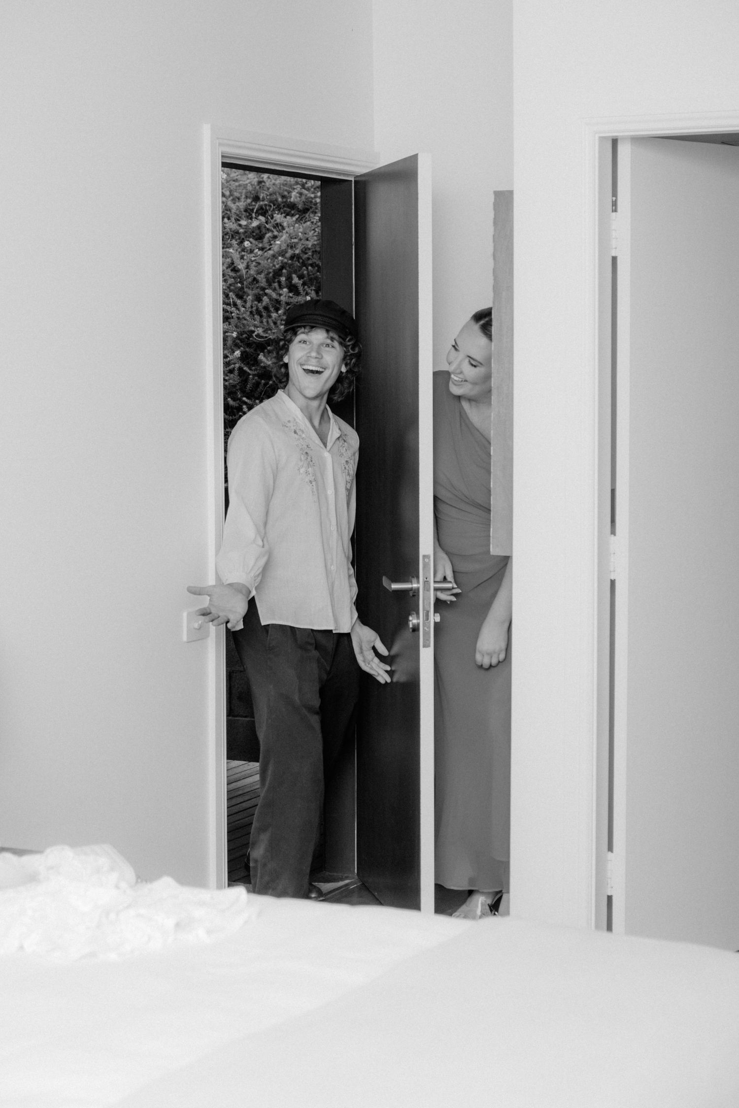 A laughing groom enters a bright doorway accompanied by a smiling woman in a black and white wedding photo.