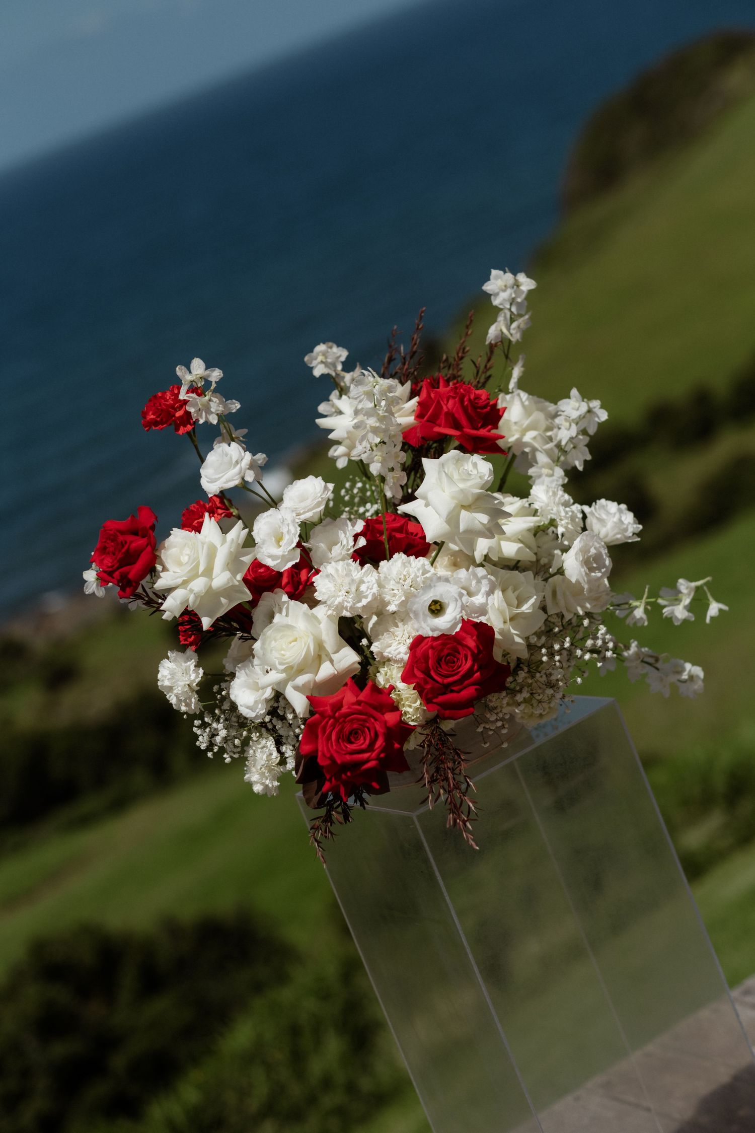 Red and white rose wedding bouquet arrangement on glass stand with ocean and green hills backdrop.