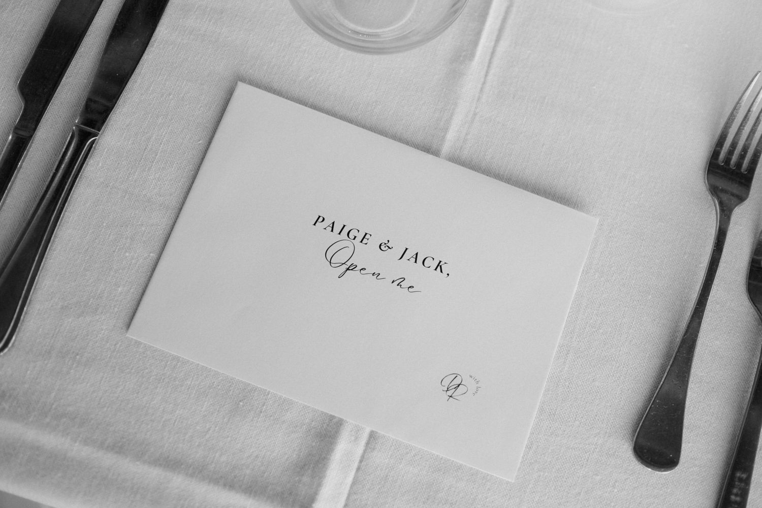 Black and white photo of a wedding place card reading Paige & Jack, Open Me, set on a white linen tablecloth.