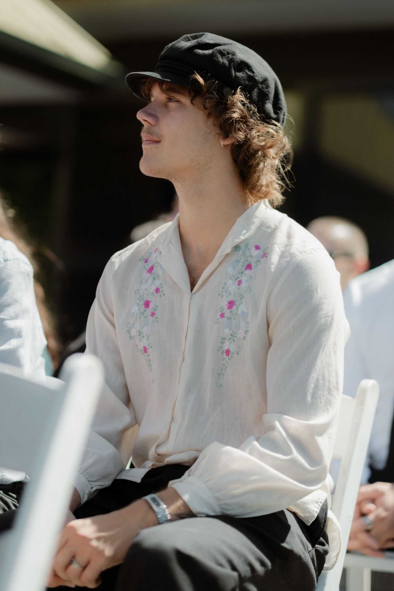 Young man in embroidered white linen shirt and black cap seated at outdoor wedding ceremony.