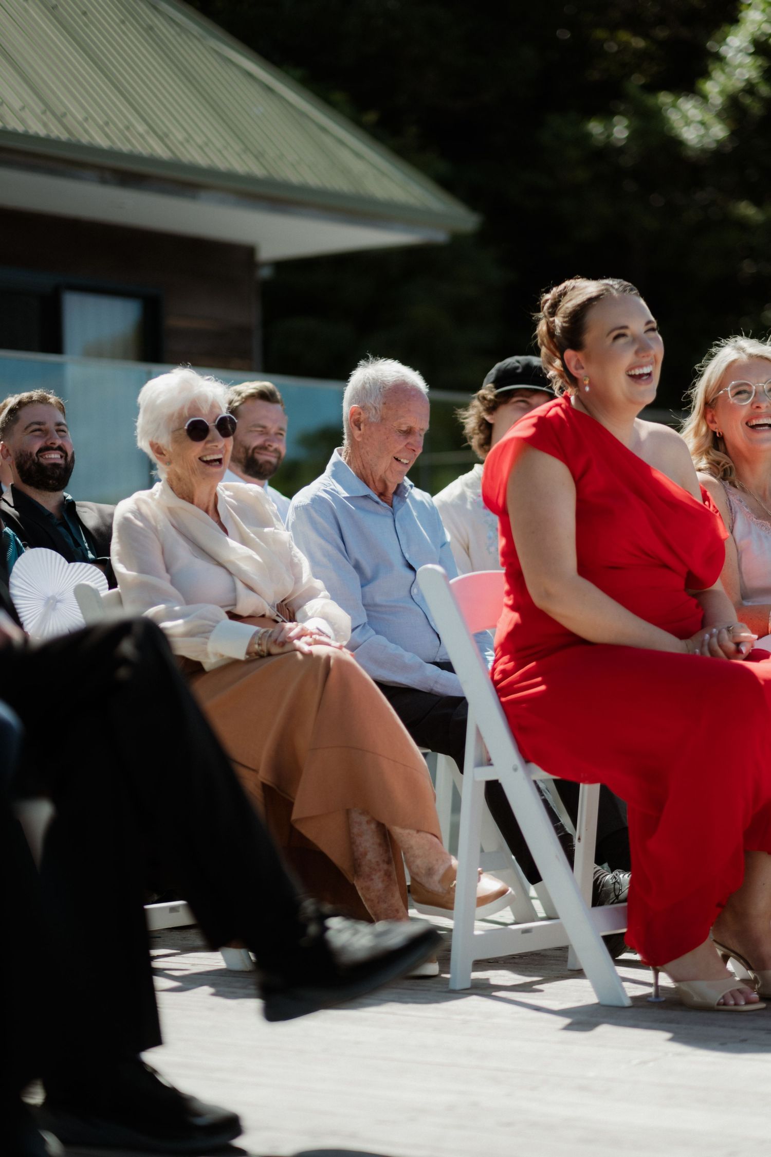 Wedding guests laughing and smiling seated in white chairs at sunny outdoor ceremony.