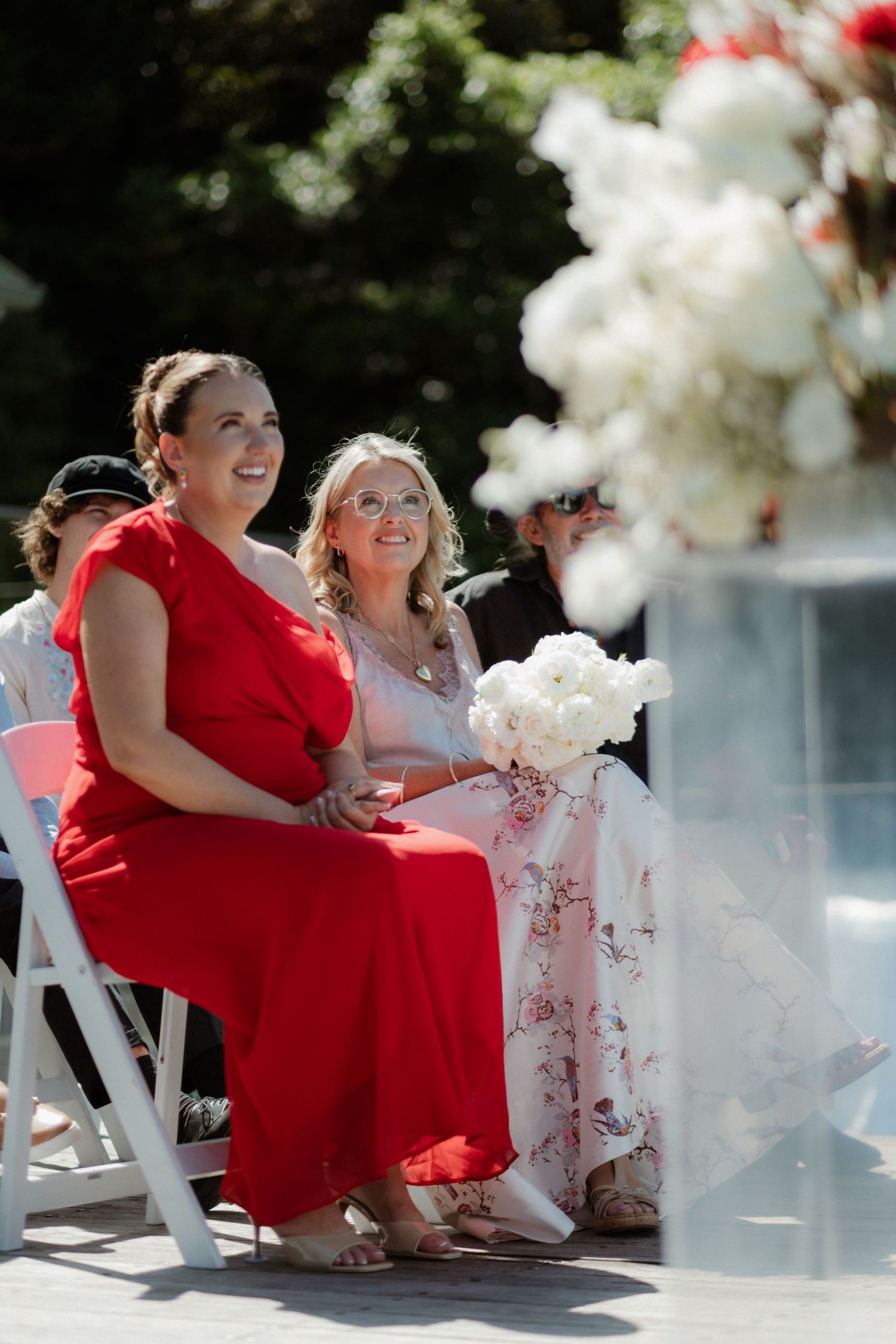 Two women in red and floral dresses seated at an outdoor wedding ceremony.