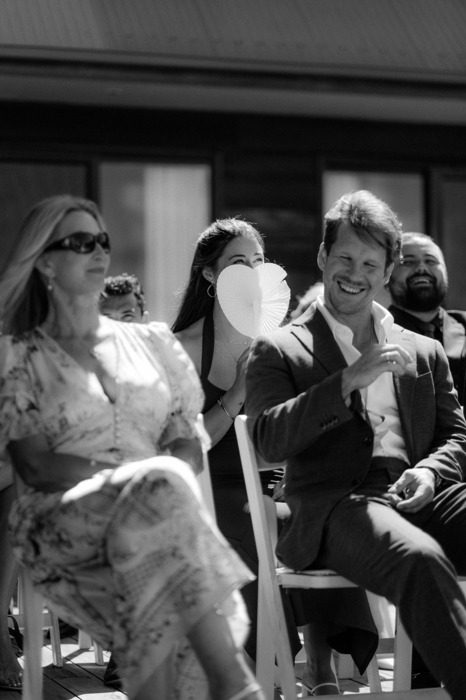 Wedding guests laugh and fan themselves in black and white photo during outdoor ceremony on a warm sunny day.