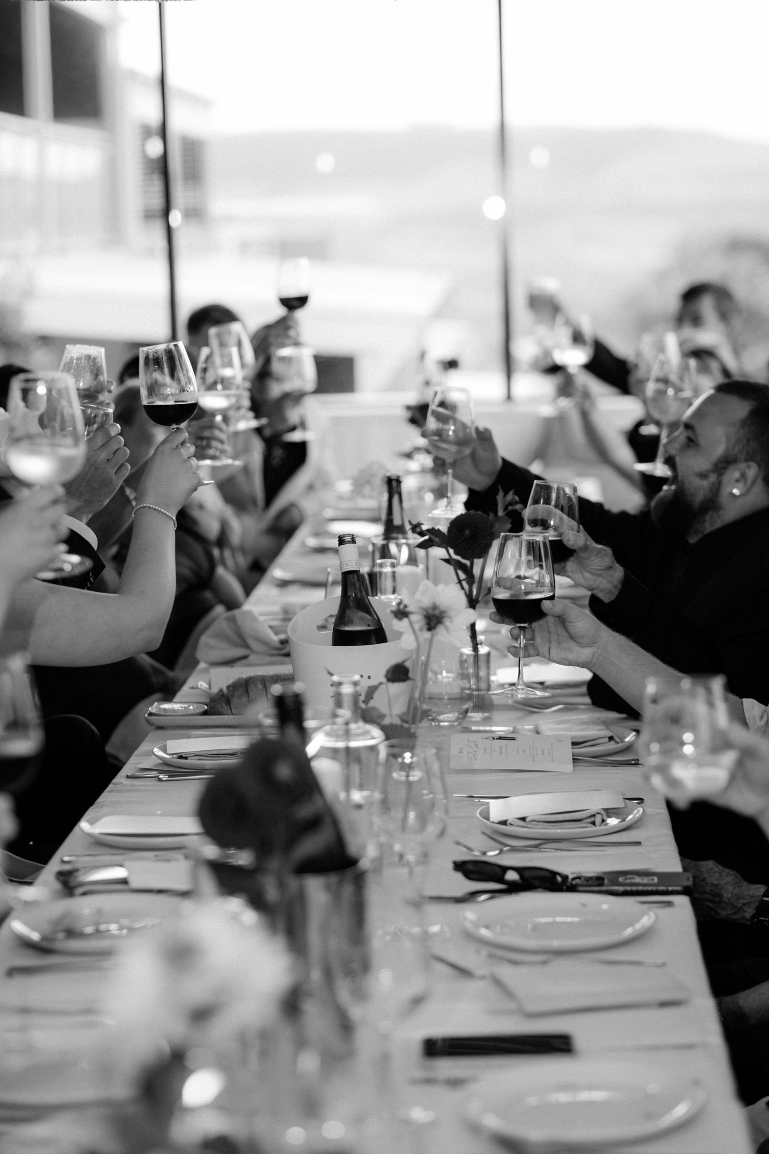Black and white photo of wedding guests raising wine glasses in a toast at a long dinner table.