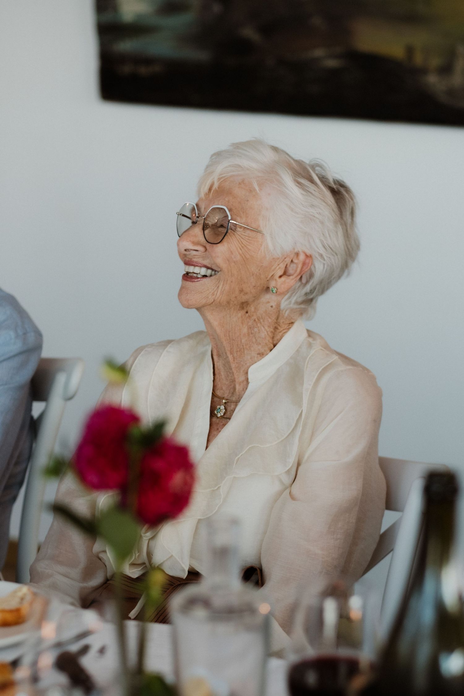 Elderly woman with white hair and round glasses laughing joyfully at a wedding reception dinner table.