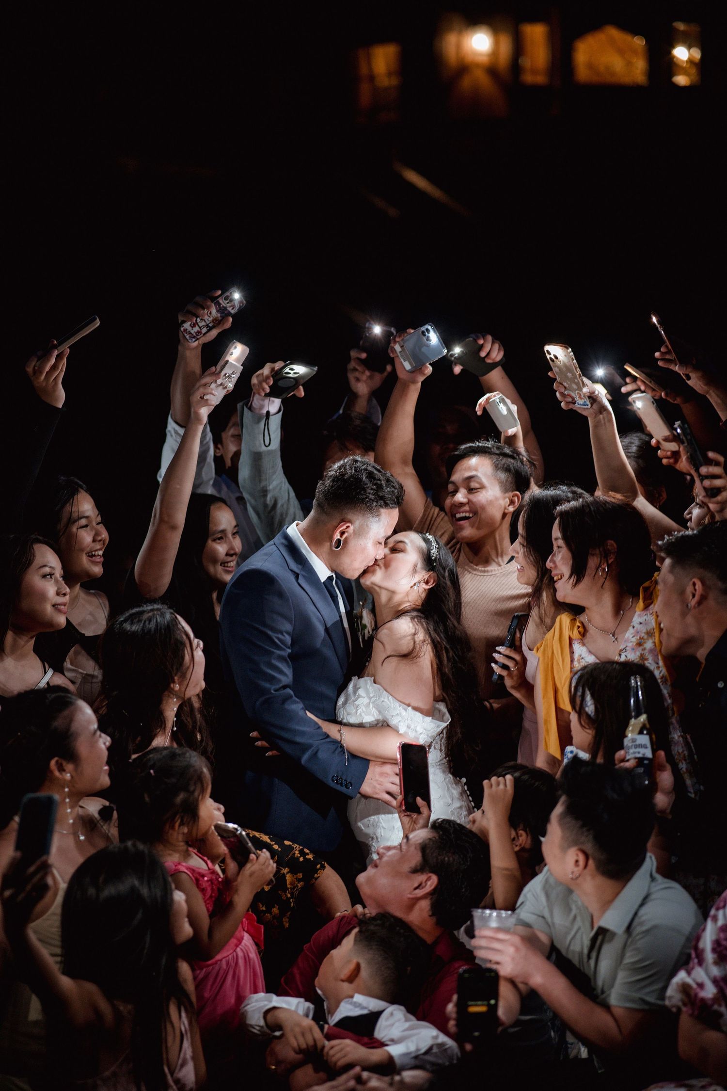 Couple shares a kiss surrounded by cheering wedding guests holding up phone flashlights in a dark reception venue.