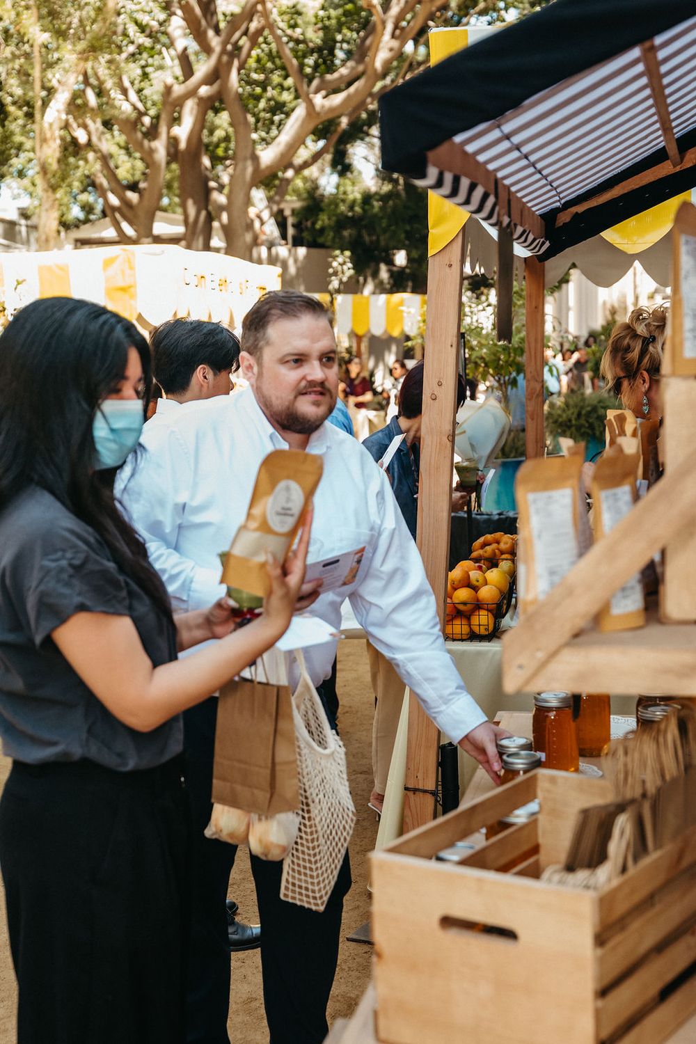 Man and woman in face mask shopping at outdoor farmers market stall with produce and packaged goods.