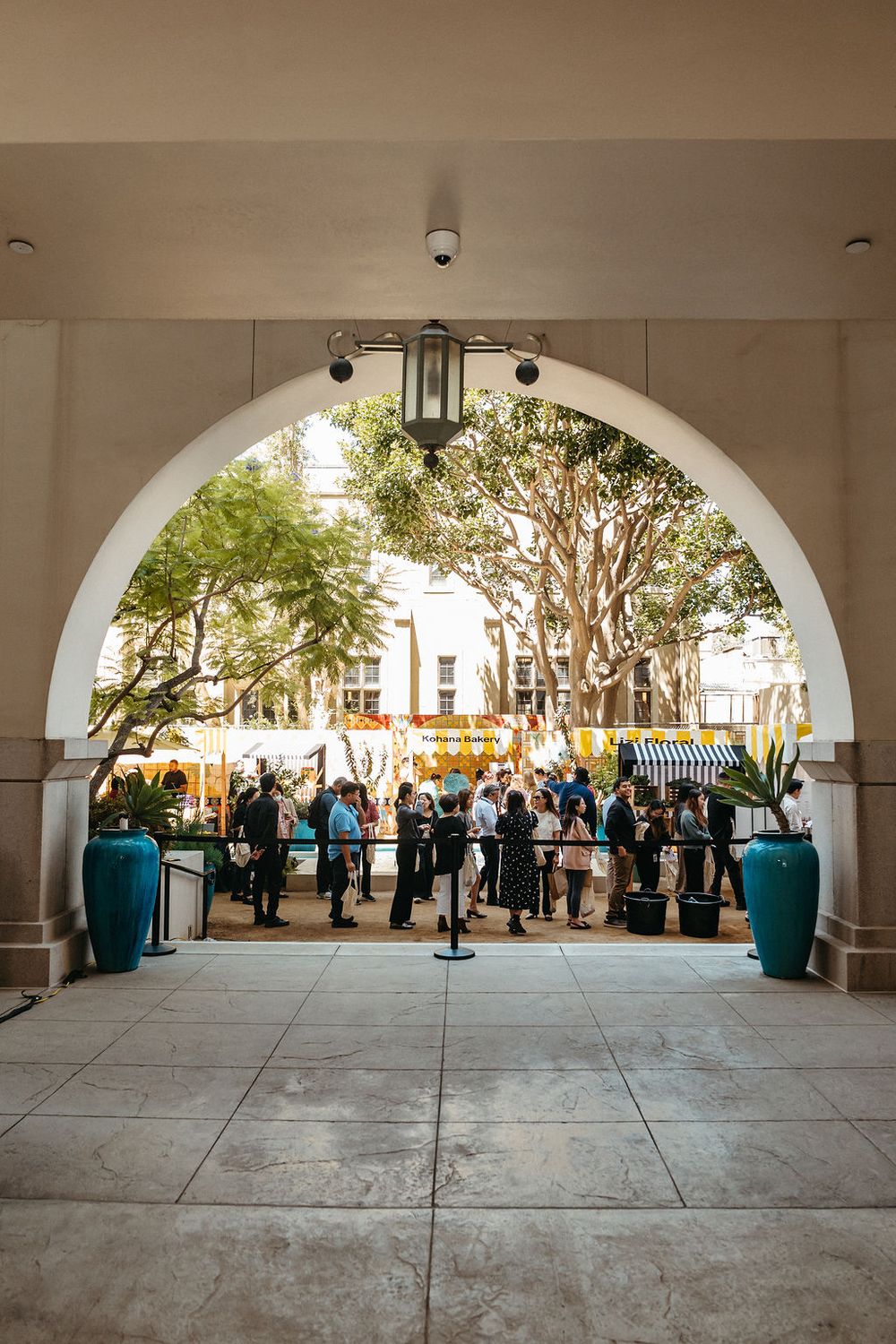 People gathered under an arched entryway at an outdoor event with lush trees and colorful market stalls in the background.