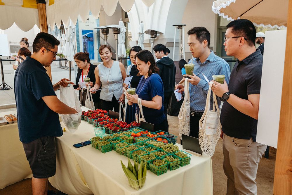 People browsing fresh produce at an indoor farmers market, selecting vegetables and fruits from a white table.
