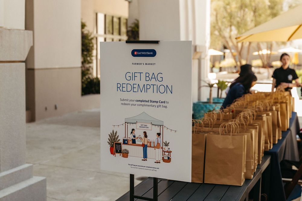 Gift bag redemption sign displayed on table with brown paper bags at outdoor corporate event.